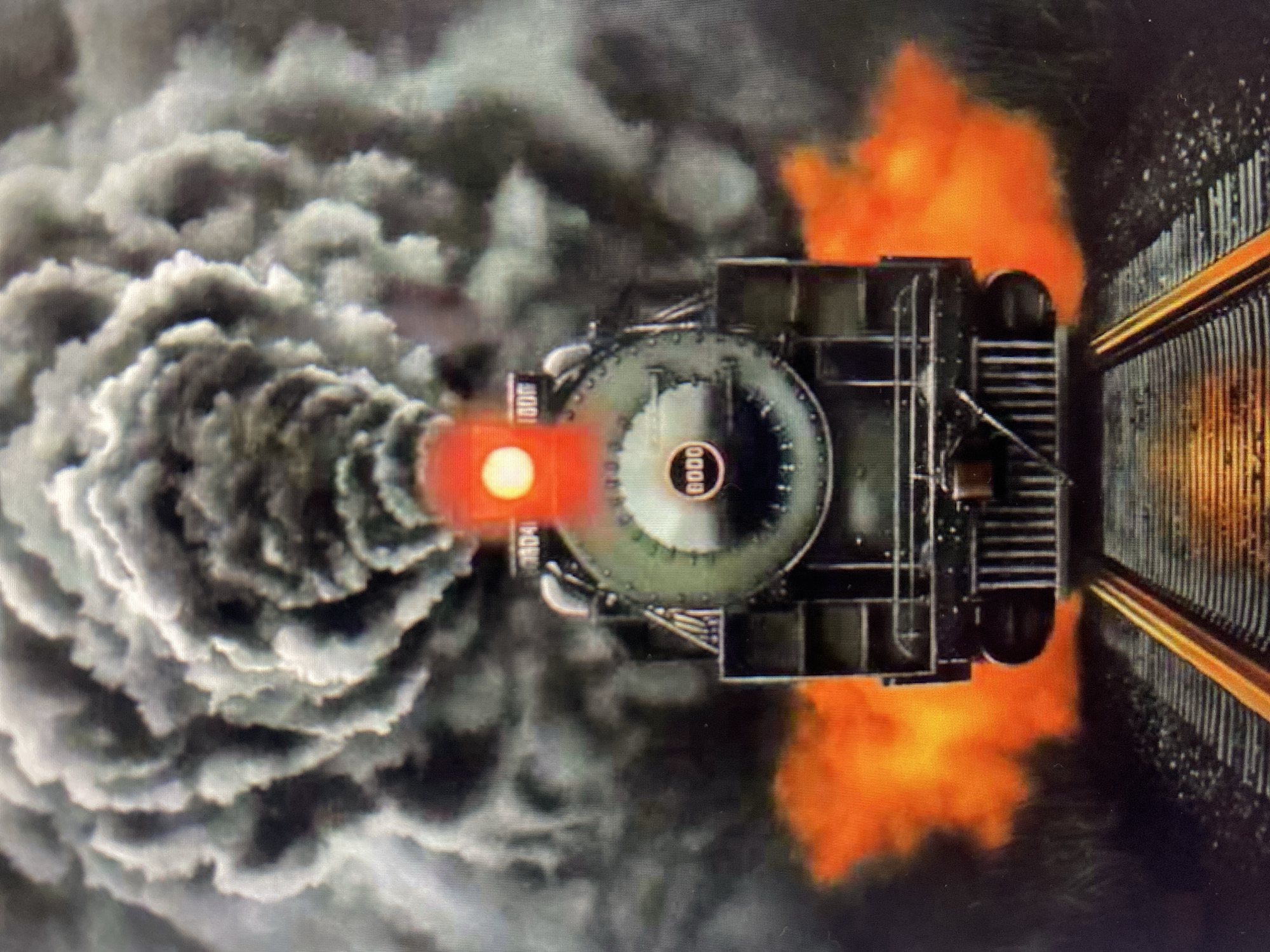 A dramatic, low-angle shot captures the front of a dark, vintage steam locomotive as it billows thick, dark smoke. The train is positioned on a set of railroad tracks that recede into the distance. Embers or flames of an orange-red hue erupt from the sides of the locomotive, creating a fiery effect against the smoky backdrop. A bright red light is visible above the train's headlight. The overall scene suggests a powerful and perhaps menacing depiction of the train in motion, possibly an artistic rendering or a scene from a film. The location is not specified in the image.