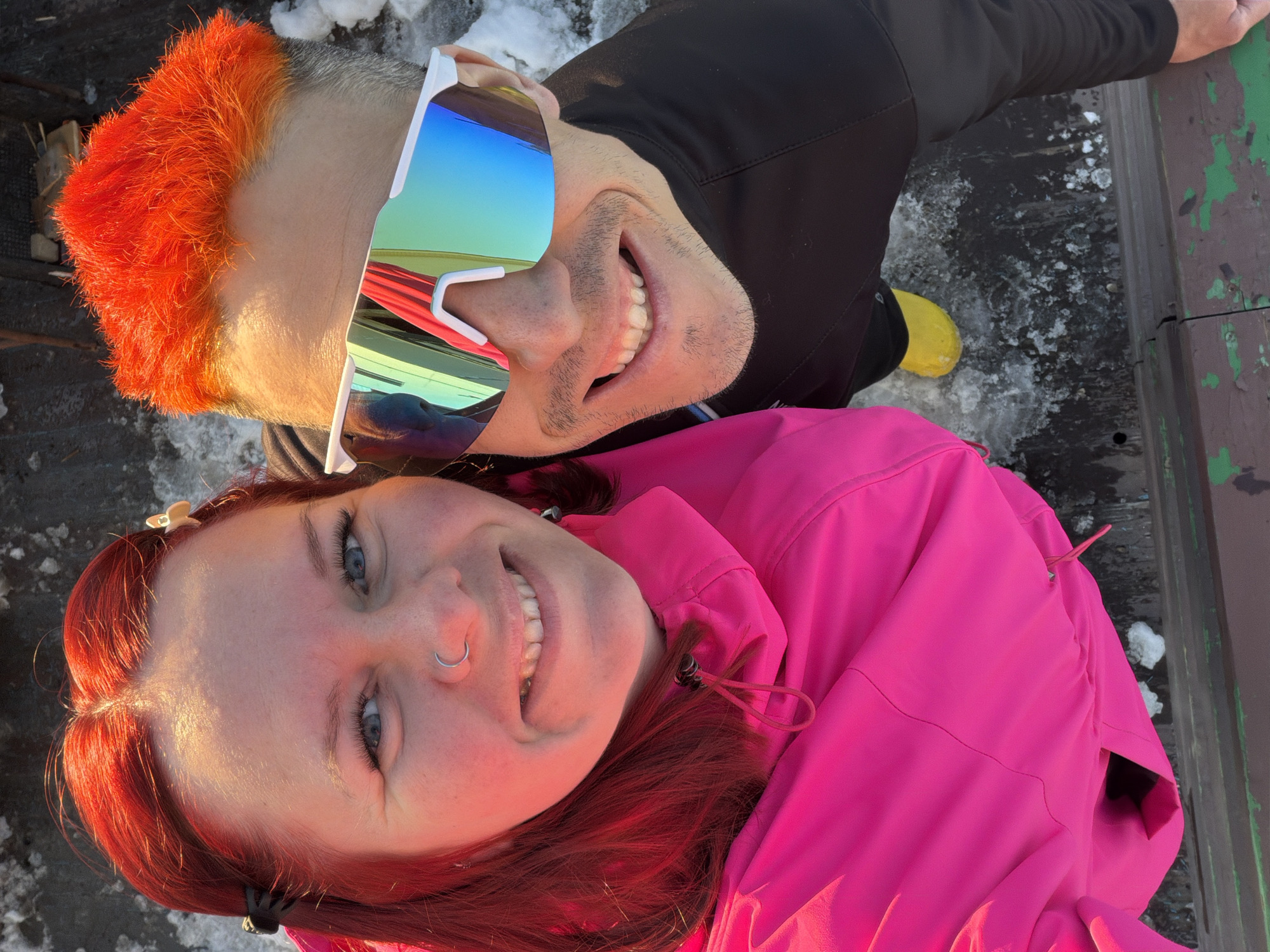 A selfie captures two individuals lying on a surface with patches of snow and textured wood. The lighting suggests daylight.
The upper figure has short, bright orange hair and wears sunglasses with reflective lenses. The person is wearing a black long-sleeved shirt. One yellow boot is visible.
The lower figure has reddish-brown hair and a nose ring. The person is wearing a bright pink jacket.
The setting appears to be outdoors, with evidence of wood and snow. The location cannot be determined from the image.