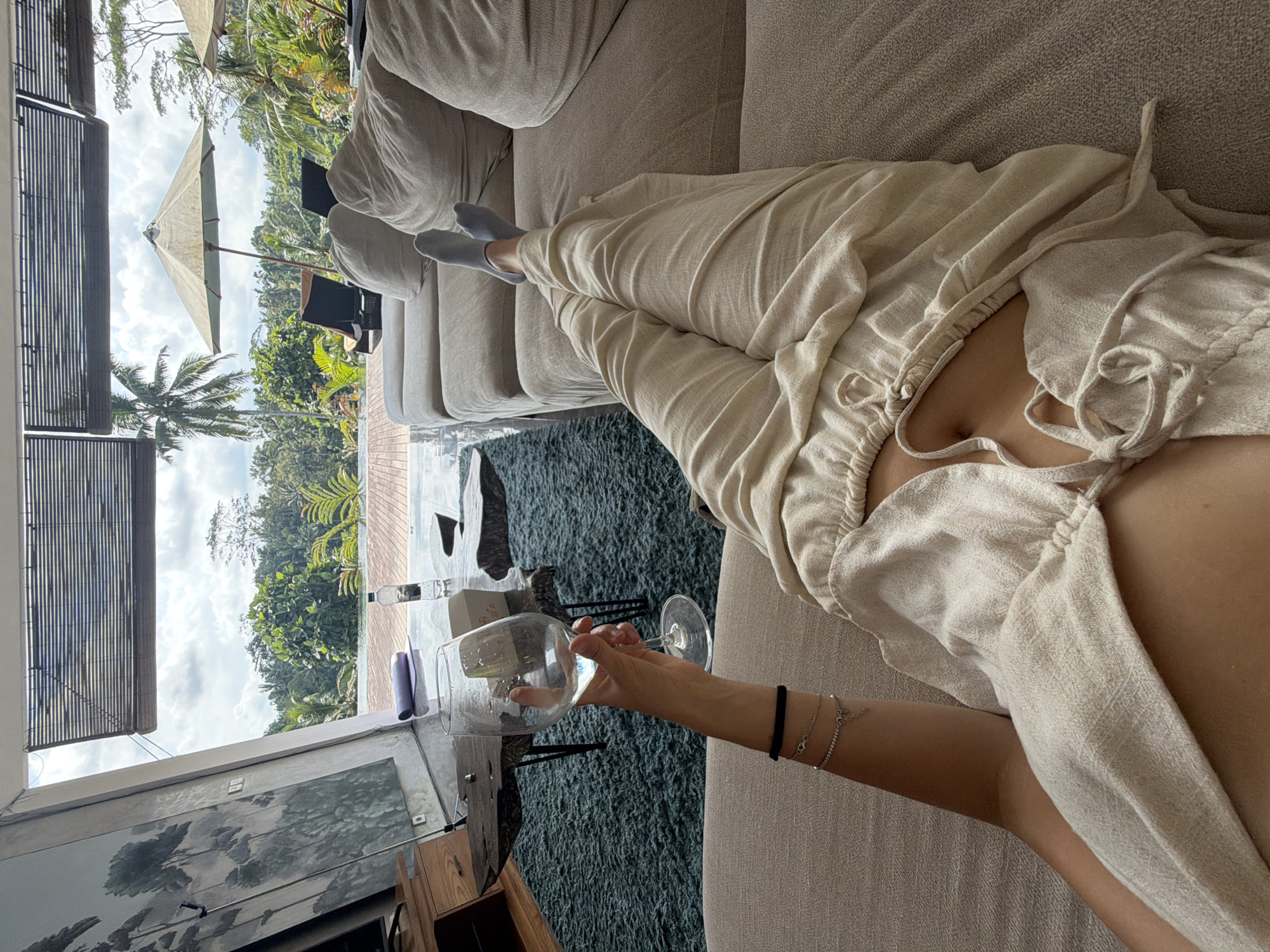A person is lounging on a beige sofa, wearing beige clothing and grey socks, and holding a partially filled wine glass. The scene is inside a building with a large window overlooking a green, tropical landscape with trees, including a palm tree, and a structure with a tan-colored umbrella. The window also shows a view of the sky with clouds. The location is likely Petulu, Indonesia, suggested by the tropical setting. The person's arm is visible, adorned with a black band and two silver bracelets. The scene includes furniture, wine glass, a person, a window, and an outdoor landscape.