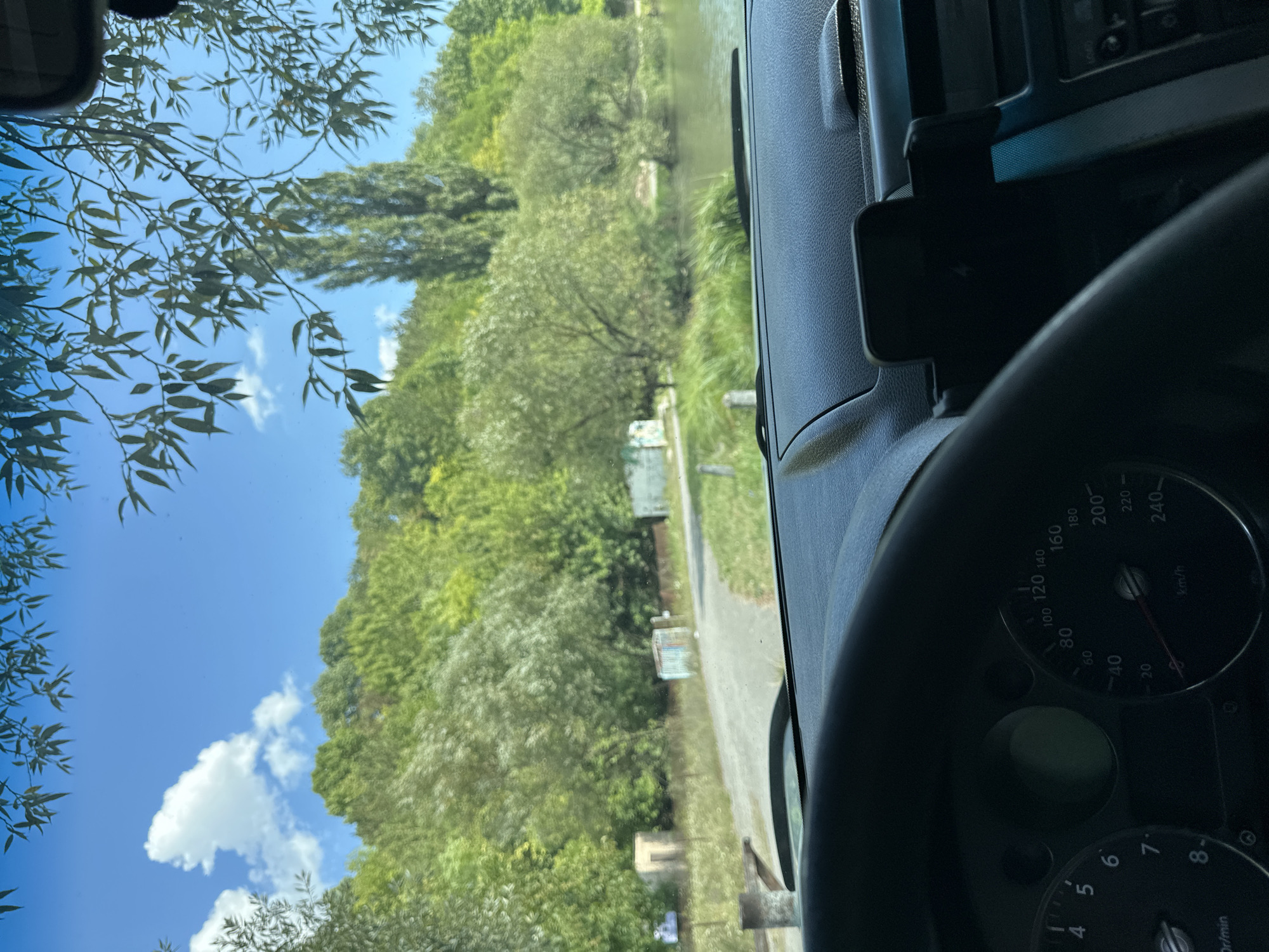 The image is taken from inside a car, looking out through the windshield at a scenic natural landscape. The foreground shows the dashboard and steering wheel of the vehicle. The dashboard includes a speedometer indicating a speed of 60 km/h and a tachometer. To the right of the steering wheel, there's a phone mount. The view outside the windshield reveals a clear blue sky with scattered white clouds. Lush green trees and vegetation cover a hillside that slopes down towards a body of water, likely a lake or river. A dirt road or path is visible in the middle ground, leading towards some small structures. The overall impression is that of a peaceful, natural setting, possibly a park or rural area near Makiv, Ukraine.
