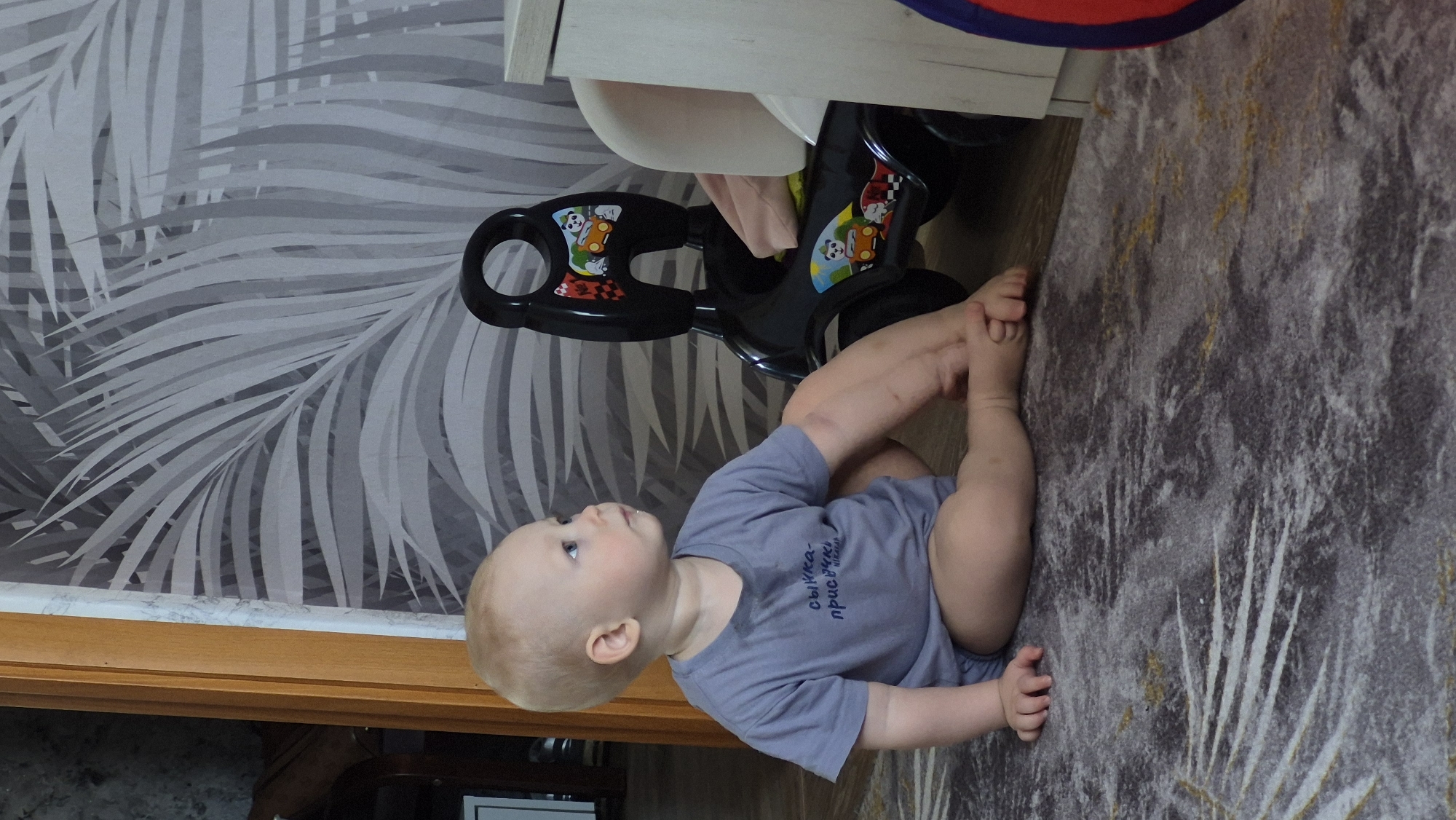 A baby wearing a gray shirt with text in Russian lies on a gray patterned rug in a room with wallpaper featuring a palm leaf design. The baby's legs are positioned upwards, with the feet near the face. A black and white walker toy is situated near a light-colored piece of furniture. Part of a wooden door frame is visible in the lower-left corner. The image was likely taken in a domestic interior in Chaikovsky, Russia.