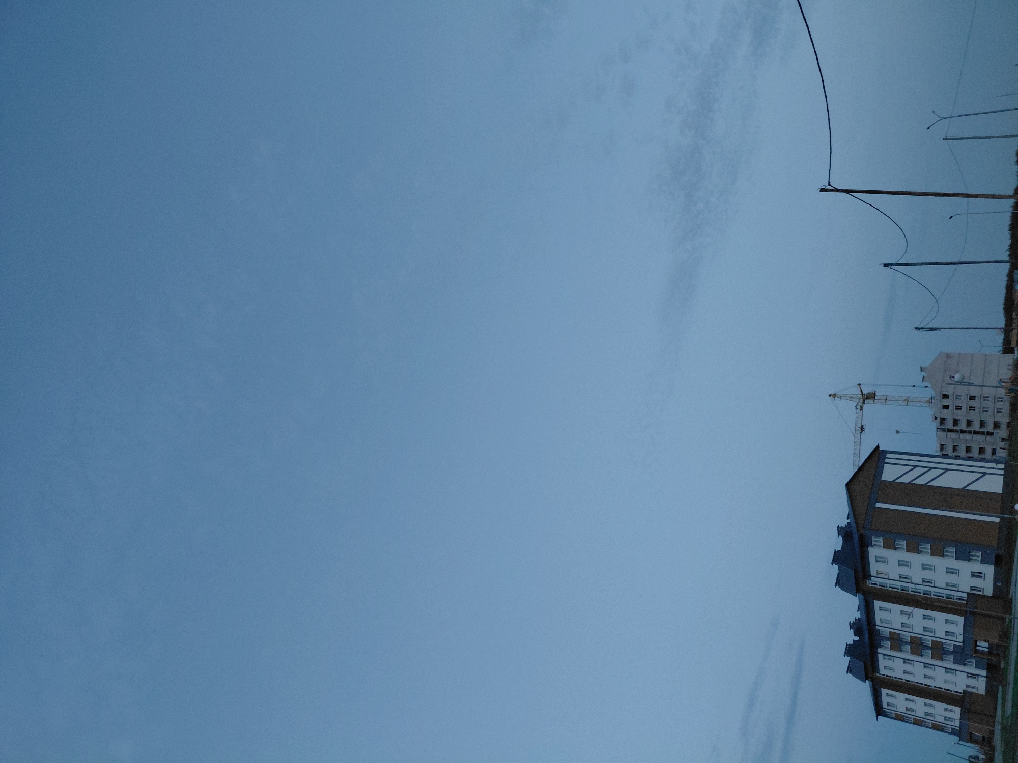 In Smarhon, Belarus, this image captures an overcast sky above a construction site. In the foreground, a cluster of multi-story apartment buildings is visible. One building, prominently featured, has a white facade with dark blue and brown accents, and a dark roof. To its right, another, less complete building is under construction. A yellow construction crane stands behind these buildings. The sky is a pale, uniform blue, with faint wispy clouds. A series of utility poles with wires stretches across the middle ground.