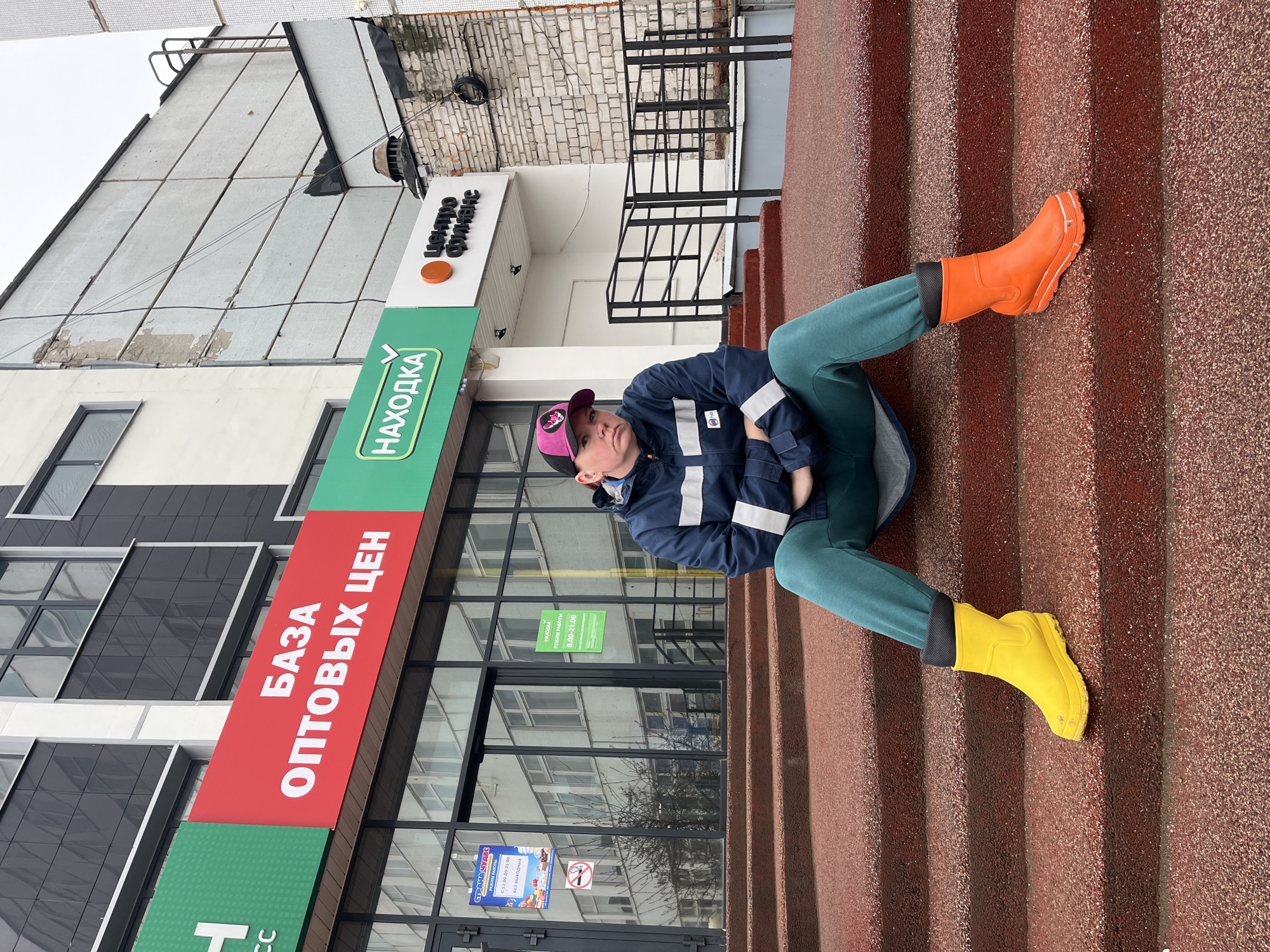 A person wearing a navy blue jacket with white stripes on the sleeves, teal sweatpants, and mismatched boots (one yellow and one orange) is sitting on red steps outside a building. The person is wearing a baseball cap with a pink brim. The building has signage in Russian that reads "БАЗА ОПТОВЫХ ЦЕН" (Wholesale Base Prices) and "НАХОДКА" (Nakodka). Additional signage states "центро финанс" (financial center) and the hours of operation. A smaller sign is visible indicating "СТРАНА ЧУДЕС" (Wonderland) and the hours of operation "С 12:00 ДО 21:00" (from 12:00 to 21:00), indicating it is open every day, also known as "БЕЗ ВЫХОДНЫХ". The photo was taken in Chaikovsky, Russia.