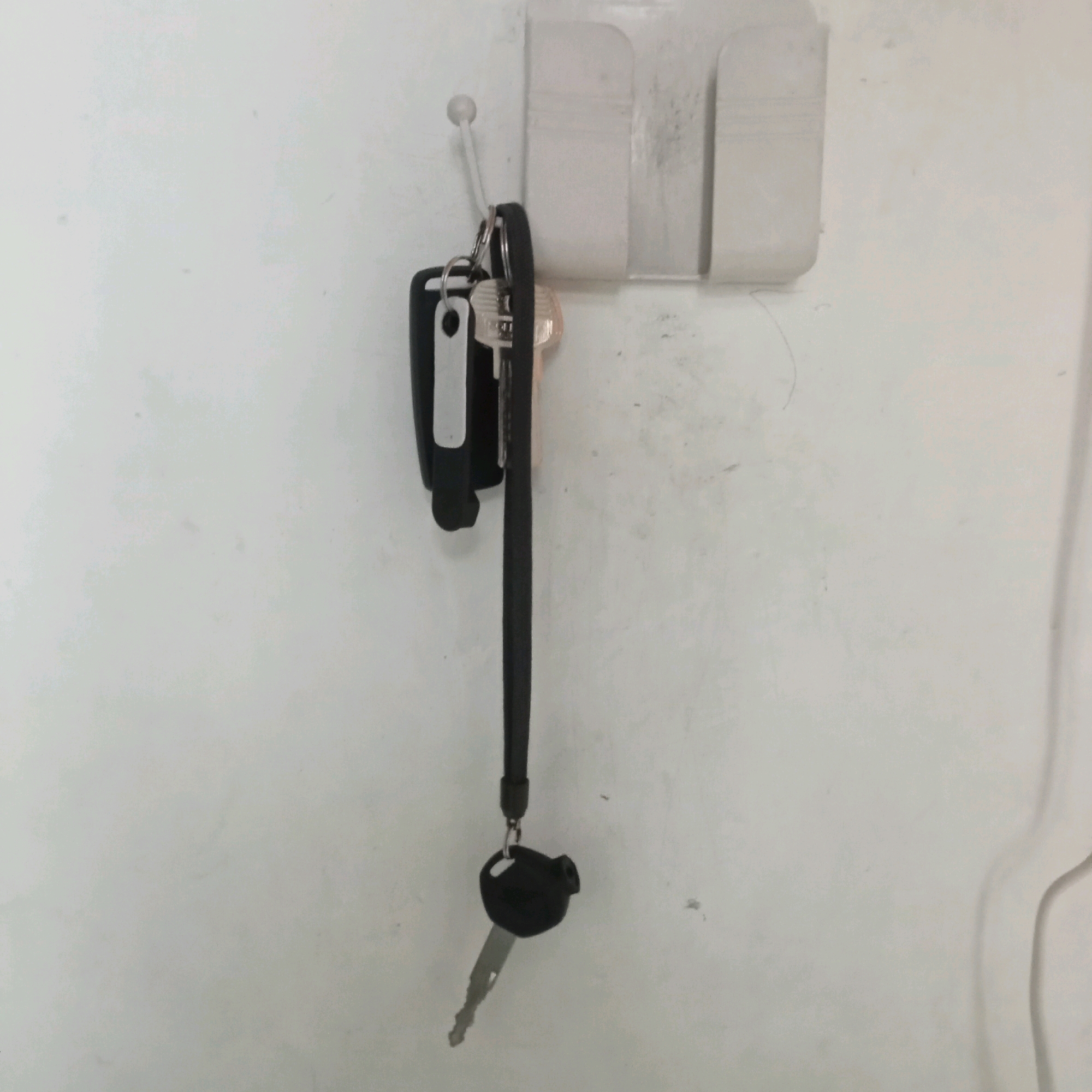 In Jaten, Indonesia, on a white wall, a transparent plastic holder with a hook is attached. A set of keys hangs from the hook, with one key featuring a black plastic fob and a silver metal blade. Another key, partially obscured, also has a black fob. A black lanyard connects the keys. The overall scene is indoors, with the wall appearing to be a neutral background.