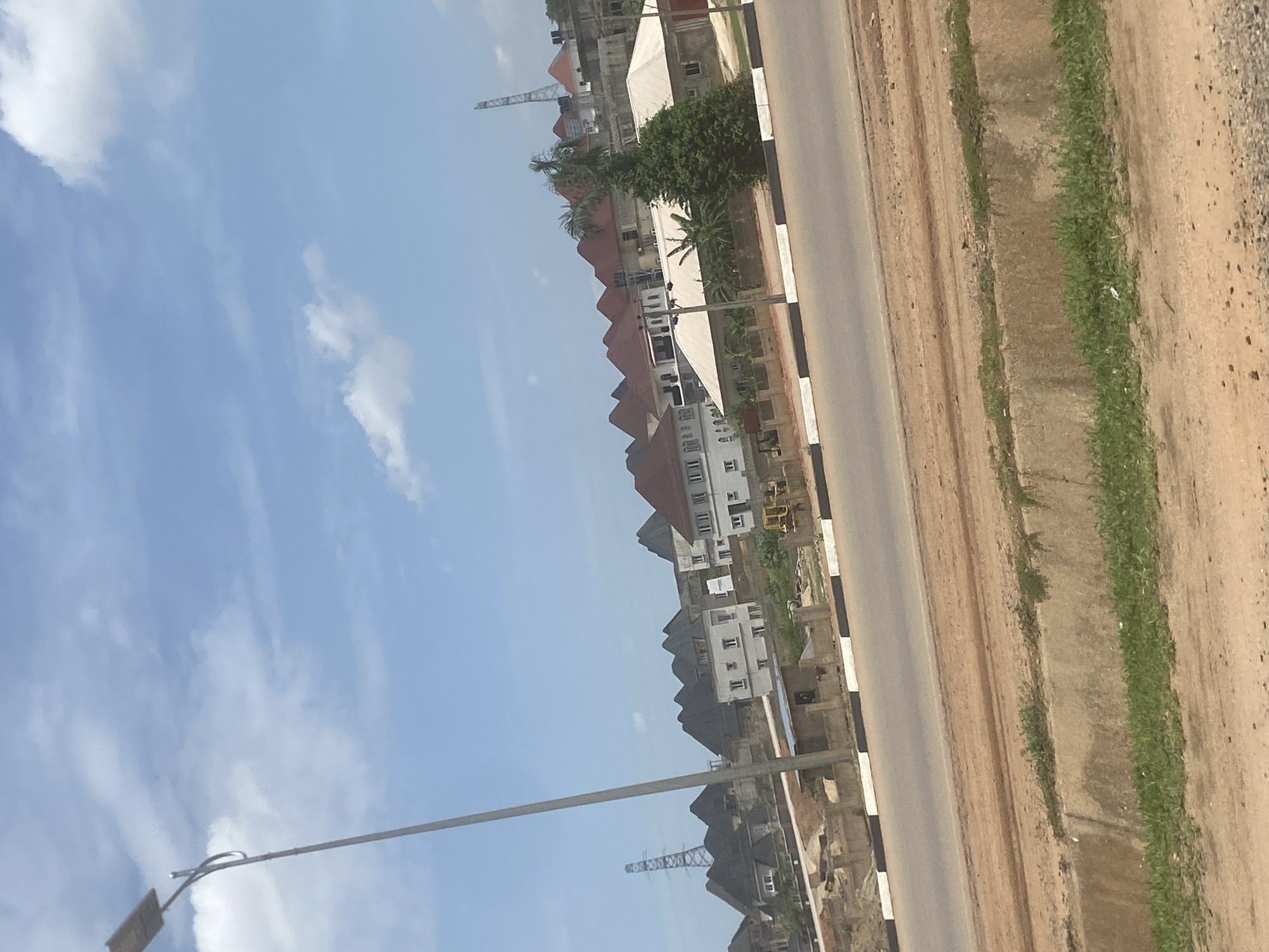 The image shows a street scene in Abuja, Nigeria. The sky is blue with some scattered clouds. A multi-lane road with a black and white curb divider runs along the right side of the image. Along the other side of the road are buildings and construction sites. A crane is visible in the distance, indicating ongoing development. The foreground includes unpaved terrain with patches of green vegetation. A light pole is visible in the lower left corner.