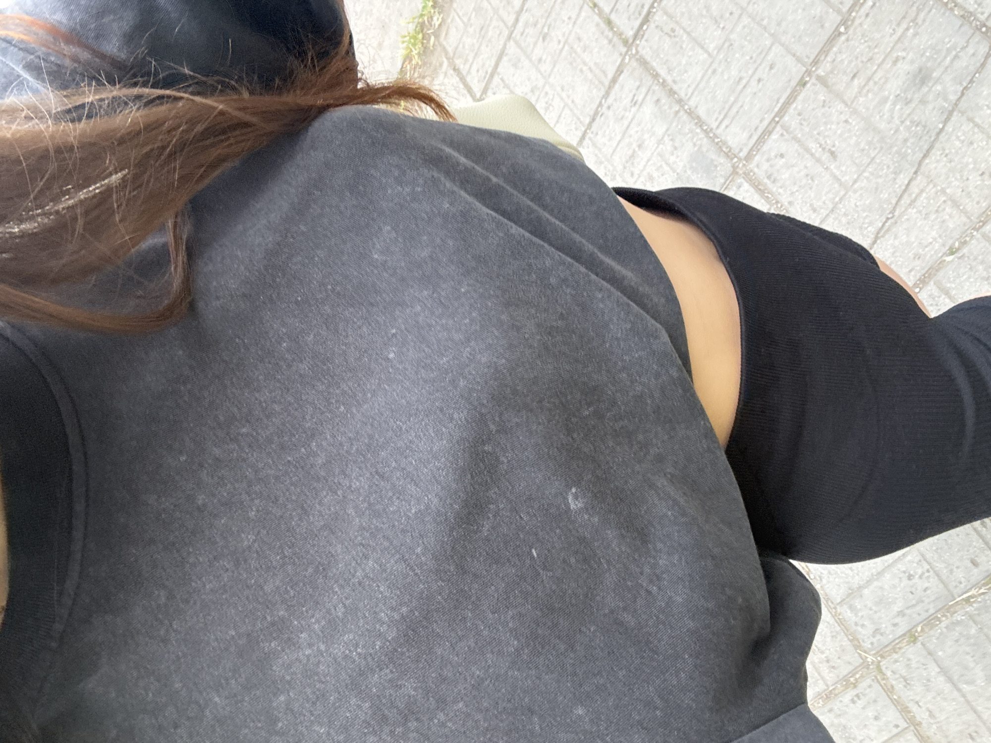 This is a close-up, waist-up shot of a person wearing a dark gray, cropped t-shirt and a black, ribbed skirt. The person has long, auburn hair that is partially visible. A light green bag is peeking out from under the t-shirt. The scene appears to be outdoors on a paved walkway with visible tile patterns. The lighting suggests it is daytime. There are no discernible landmarks to pinpoint a specific location within Sofia, Bulgaria.