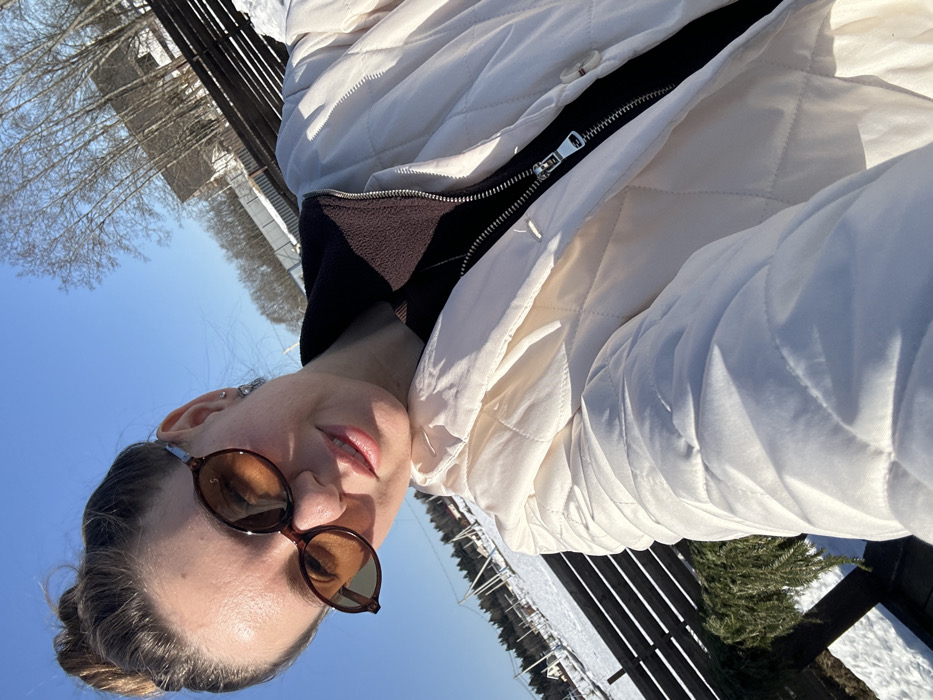 A female individual is captured in a selfie on a bright, sunny day in a snowy, outdoor environment identified as Shipulino, Russia. She wears dark brown, oval-shaped sunglasses, a light-colored, quilted jacket (cream or off-white) over a dark inner layer with a visible zipper and brown fleece collar. Her brown hair is styled in a bun, and she has small silver-toned earrings. Her lips are tinted light pink, and she exhibits a slight smile. The background reveals patches of snow on the ground, a dark brown wooden fence running horizontally, and several bare deciduous trees. In the further distance, residential-style buildings with pitched roofs are observable. To the left, a section of netting or fencing is visible, along with a fallen evergreen branch lying on the snow-covered ground near the fence. The sky is clear and blue.