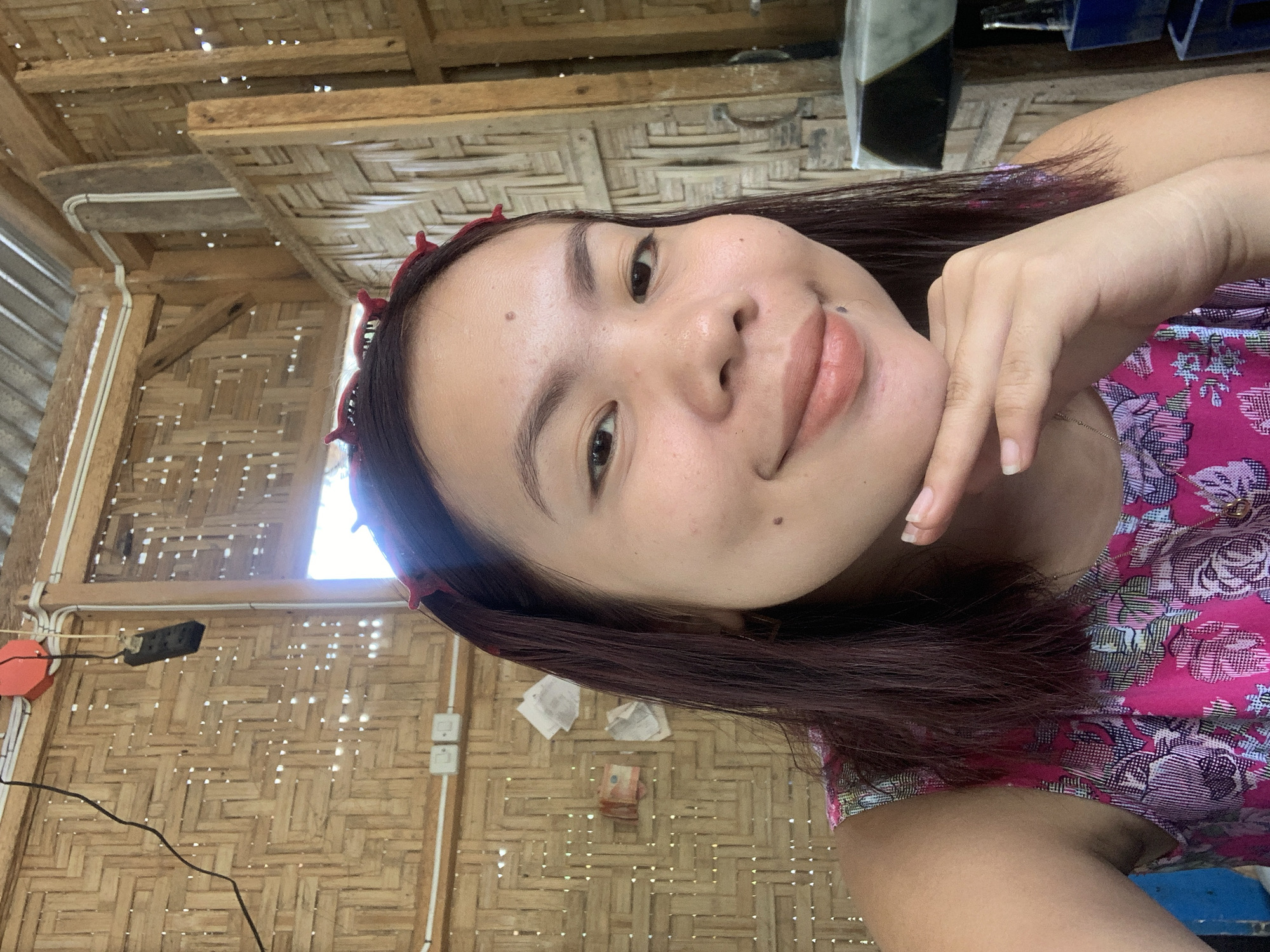 A young woman with dark hair and a red headband is captured in a close-up selfie. She is wearing a vibrant pink floral top and resting her chin on her hand, with a slight smile. Her skin has a few blemishes and a mole near her lips. The background consists of woven bamboo walls, typical of rural Philippine architecture, and a corrugated metal roof. Electrical outlets and wires are visible on the wall, along with some pieces of paper and what appear to be banknotes pinned to the bamboo. The scene suggests a simple, possibly outdoor, setting in a dwelling located in Kiblawan, Philippines.