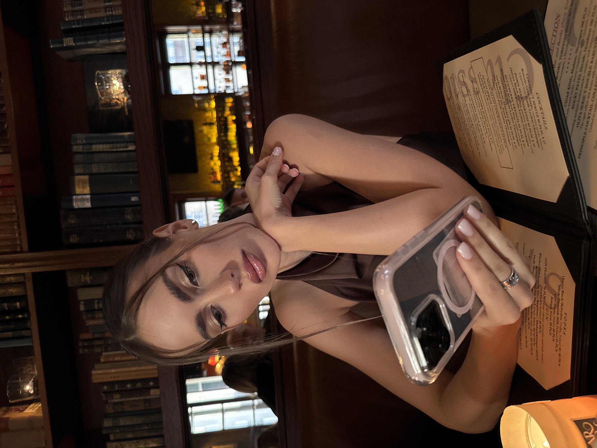 A young woman sits at a table in a dimly lit room resembling a bar or lounge, likely in Unknown. She holds a smartphone in her right hand, displaying it towards the viewer. The woman has fair skin, dark eyeliner, and pink glossy lips. She wears a sleeveless brown top. On the table next to her are two cocktail menus in black folders. Behind her, a bookshelf filled with various books is visible. A back wall has illuminated shelves containing bottles. The location appears to be a bar in an urban area.