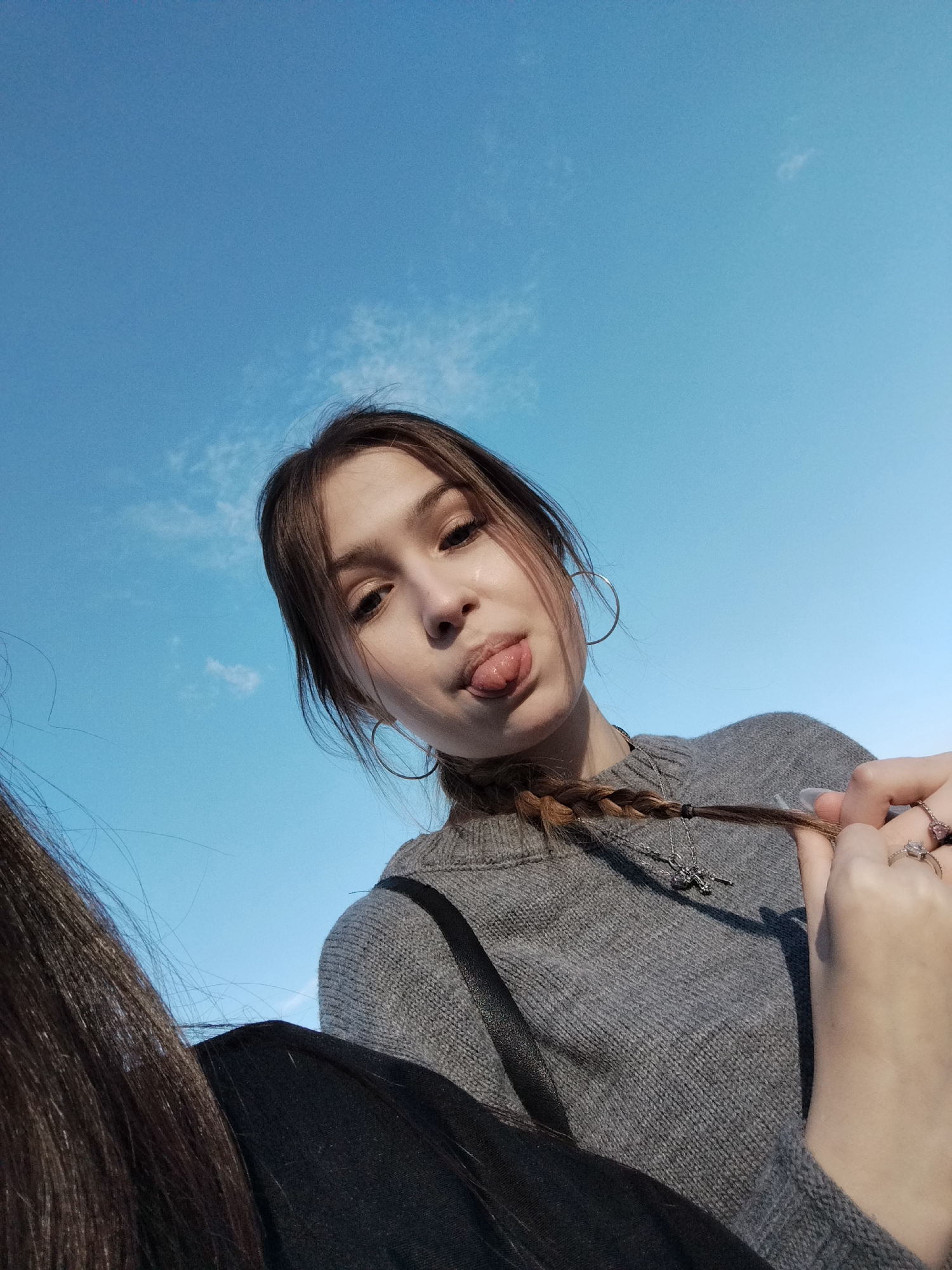 A young woman is seen from a low angle, likely a selfie, against a bright blue sky with minimal clouds. She is sticking her tongue out and looking down at the camera. She has brown hair with a braid, hoop earrings, and is wearing a gray sweater. She has a silver necklace with a cross pendant. Her right hand is visible, holding the end of her braid, with rings on her fingers. The edge of another person's hair and a black garment are visible on the left side of the frame. The location, based on the request, is presumed to be Chaikovsky, Russia, though there are no visible elements in the photo to confirm the specific location within the city.