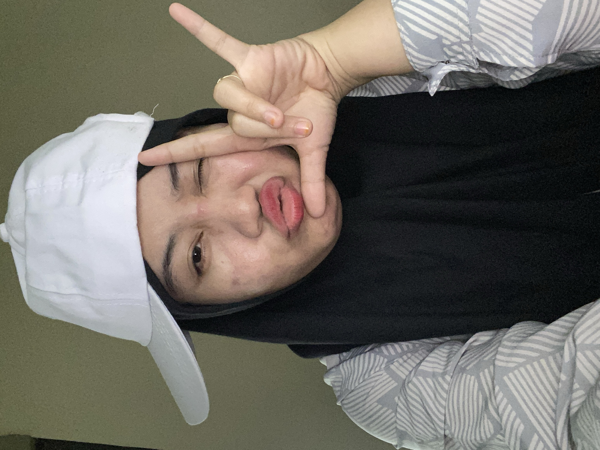 A young woman wearing a black hijab and a white baseball cap poses for a selfie. She is winking and puckering her lips, with her right hand making a "L" shape gesture. Her shirt has a gray and white striped pattern. The background is a plain, light brown wall. The image appears to be taken indoors, likely in a residential setting in Banjarbaru, Indonesia.