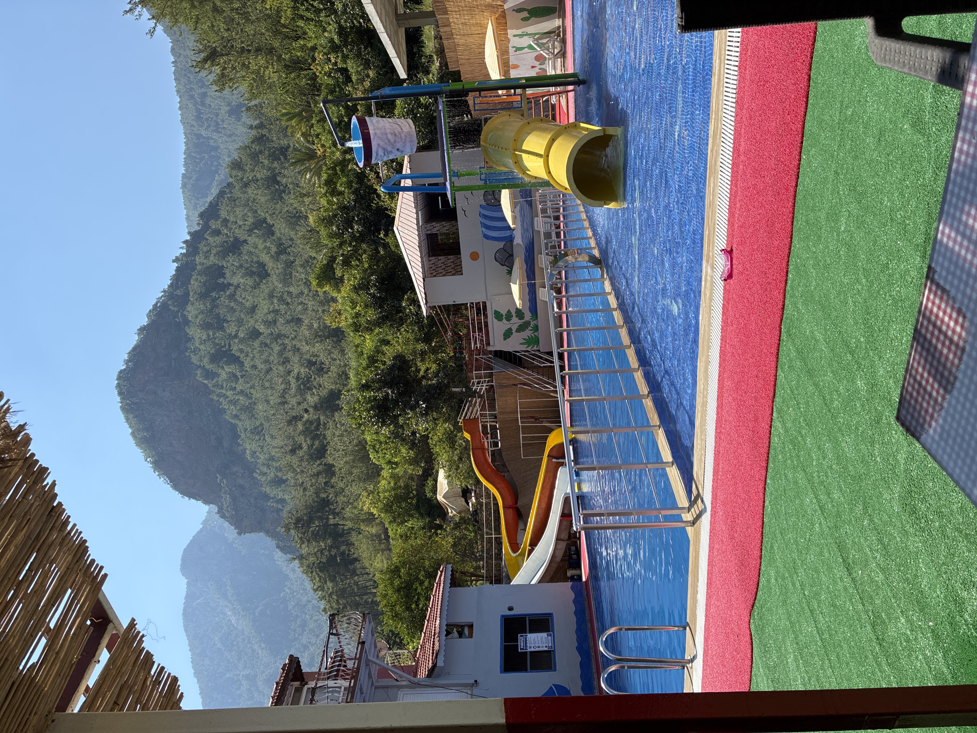 The image shows a water park in Alanya, Turkey, on a sunny day. The park features a large blue swimming pool with a tiled bottom, surrounded by a red and green artificial turf area. Several water slides are visible: a large yellow tube slide, and two other slides, one orange and one white, that merge into a common run-out. A splash bucket filled with water is suspended above the yellow tube slide. In the background, a building with a red roof and balconies is partially visible, along with lush green hills and mountains. The foreground shows a portion of a thatched roof structure and a table with a checkered tablecloth. There are no people visible in the water park itself.