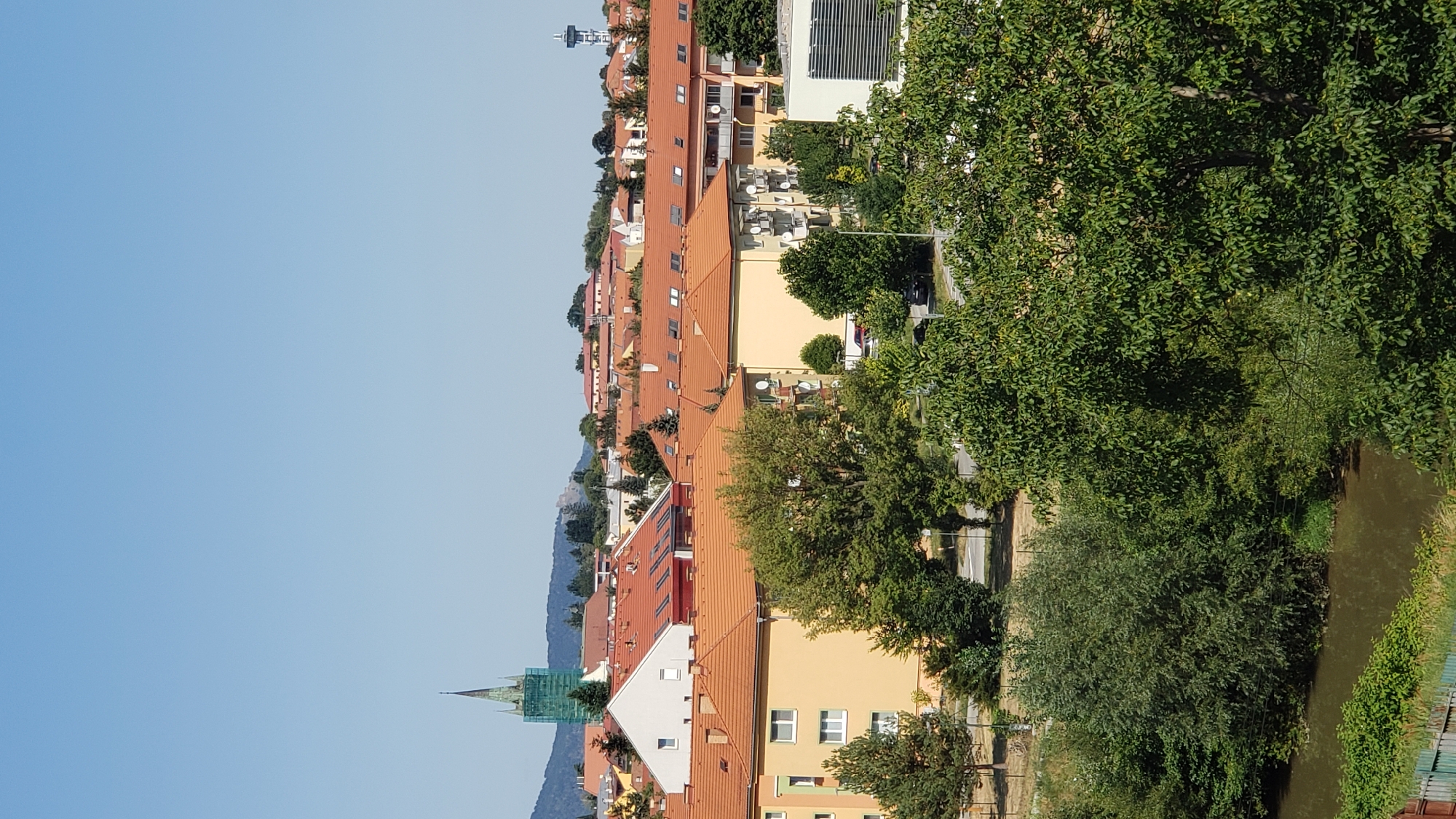 The image shows a view of a cityscape, likely in Prešov, Slovakia, under a clear blue sky. In the foreground, dense green trees and foliage dominate the lower portion, with a narrow, murky body of water visible on the far left. Behind the trees, the mid-ground features a collection of apartment buildings with orange tiled roofs, characterized by multiple windows and balconies. Some of these buildings are yellow, while others have white facades. A prominent feature in the background, slightly to the left, is a tall, slender green church tower, partially obscured by scaffolding. Further in the distance, rolling hills and other buildings can be seen, including what appears to be a tower with a radio antenna on the right side of the frame. The scene appears to be a residential area with some historical or religious structures visible. There are no people visible in the image.