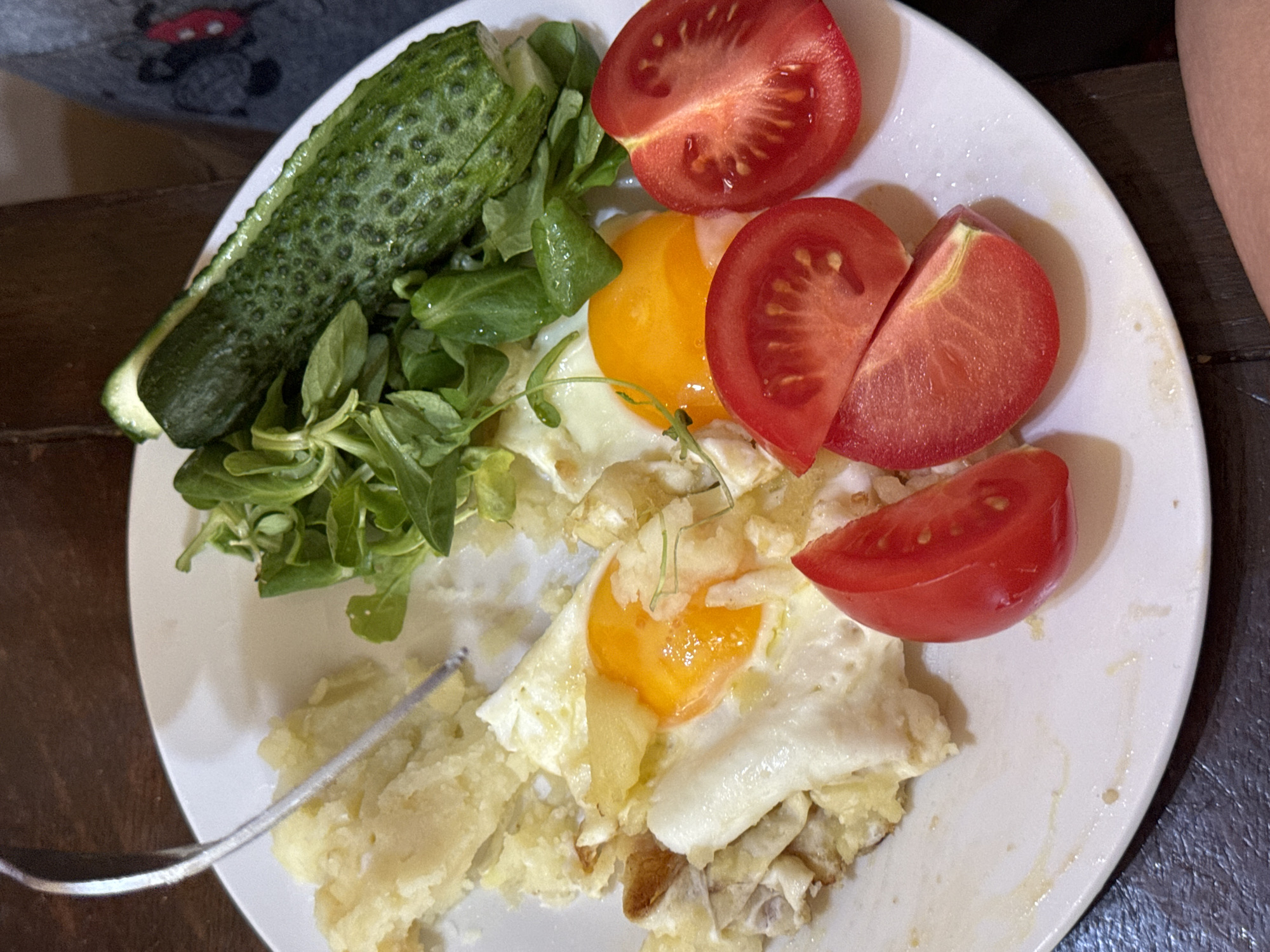 The image shows a close-up view of a white plate containing a meal. The meal consists of two fried eggs with visible yolks, mashed potatoes, sliced tomatoes, a piece of cucumber topped with greens, and some other food items that are difficult to identify clearly. A fork is visible on the left side of the plate, in contact with the mashed potatoes. The background appears to be a wooden table. There are no visible landmarks or environmental cues that would indicate a specific location in Waterford, Ireland. The context of the meal suggests it is likely indoors, possibly in a home or a casual dining setting.