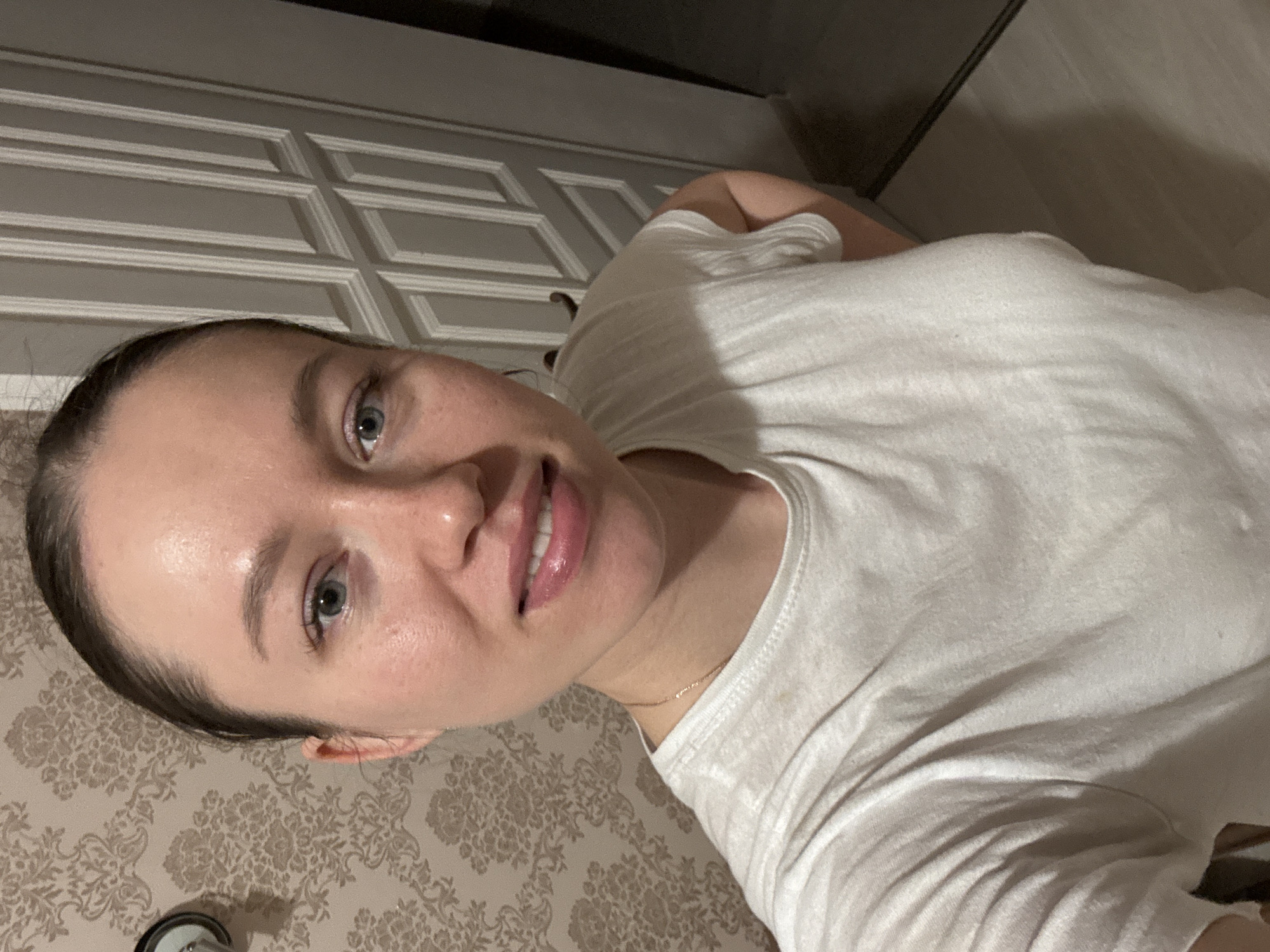 A woman with fair skin, blue eyes, and dark hair pulled back is posing for a selfie. She is wearing a white t-shirt and a thin gold necklace. The background includes a patterned beige wall and a gray door with white paneling. The location of the image is Ферма, Russia.