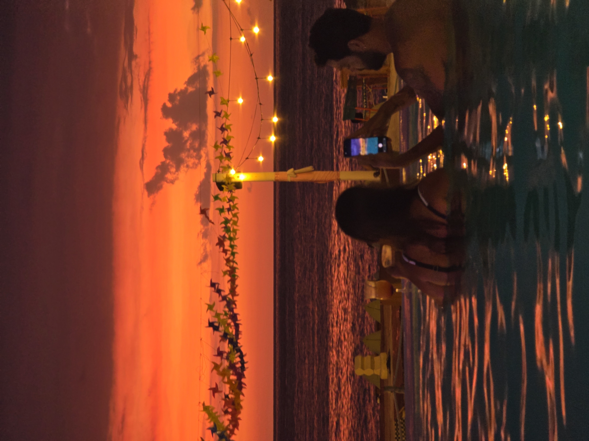 Here is a forensic description of the image:
The image depicts a sunset scene at an outdoor bar or restaurant situated by the sea. The sunset casts an orange and red hue across the sky and reflects on the water. A string of lights and colorful decorations are suspended above the scene. A man and a woman are partially submerged in what appears to be a pool or shallow water near the bar. The man is taking a picture with his phone, while the woman is holding a drink. The location is a beach bar or restaurant. The city and country are unknown.