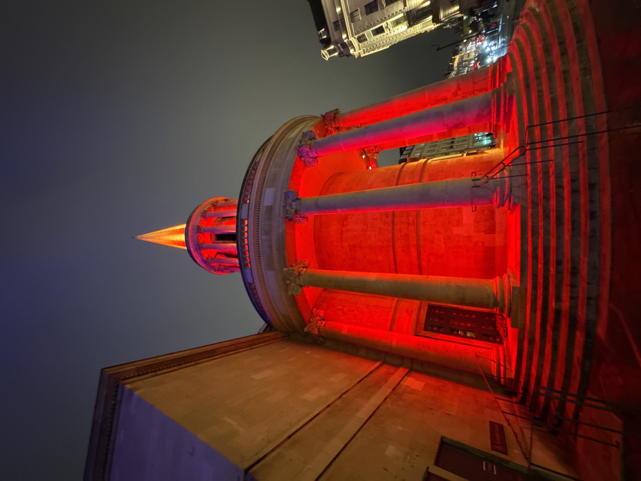 An eye-level perspective captures a building with prominent columns, illuminated in a vibrant red hue, against a darkening evening sky. A spire-like structure atop the building glows with a golden-orange light. To the right, a staircase is visible, also bathed in red light, while a neighboring building is partially seen in the upper portion of the image.