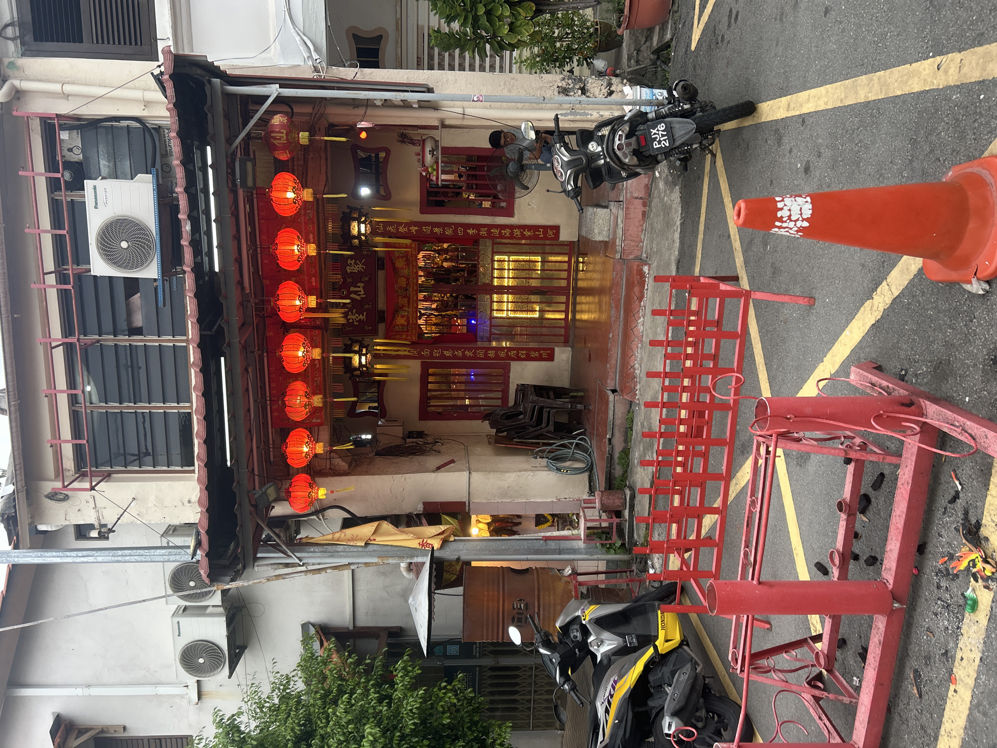 The image depicts a narrow street in George Town, Malaysia, likely in a heritage area based on the architecture. The focal point is a brightly lit storefront, possibly a temple or a shop with religious significance, adorned with numerous red lanterns. The building features a red-tiled roof and a facade with intricate details, including Chinese calligraphy above the entrance. Two motorcycles are parked in front of the building, one is yellow and grey, the other is black. A man wearing glasses and a grey t-shirt is seated on the black motorcycle.
In the foreground, a red metal structure with circular holders is placed on the asphalt, surrounded by scattered debris, possibly remnants of fireworks. Another red metal barrier is visible behind it. An orange traffic cone with white markings stands near the motorcycles. A lush green tree is on the left side of the image, and potted plants are visible on the right. The building also has air conditioning units mounted on its exterior walls. The overall impression is a scene of urban street life with cultural elements, suggesting a festive or religious occasion.
