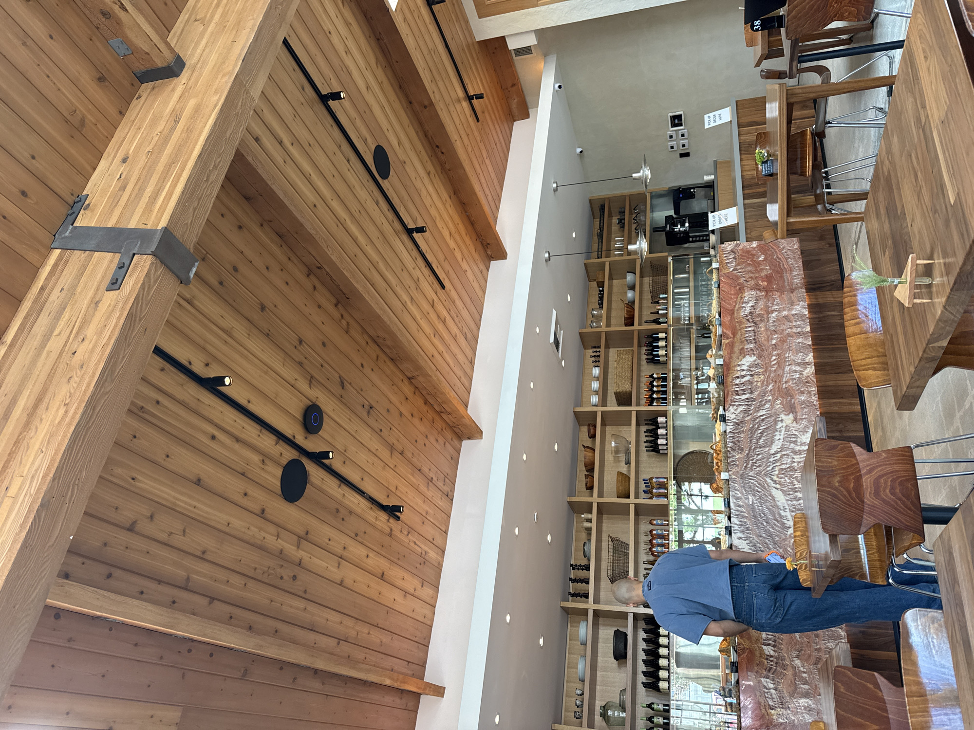 The image shows the interior of a cafe or restaurant, possibly located in Los Angeles, United States. The distinctive architectural elements include a high, pitched ceiling constructed of wood planks, reinforced with visible steel brackets and lit by track lighting. One wall is lined with built-in wooden shelves stocked with wine bottles, glassware, and decorative items. A man in a blue shirt and jeans stands near a counter made of stone or marble, possibly ordering or waiting for food. Seating includes wooden chairs and tables, some with high stools. The space has a modern, upscale feel.