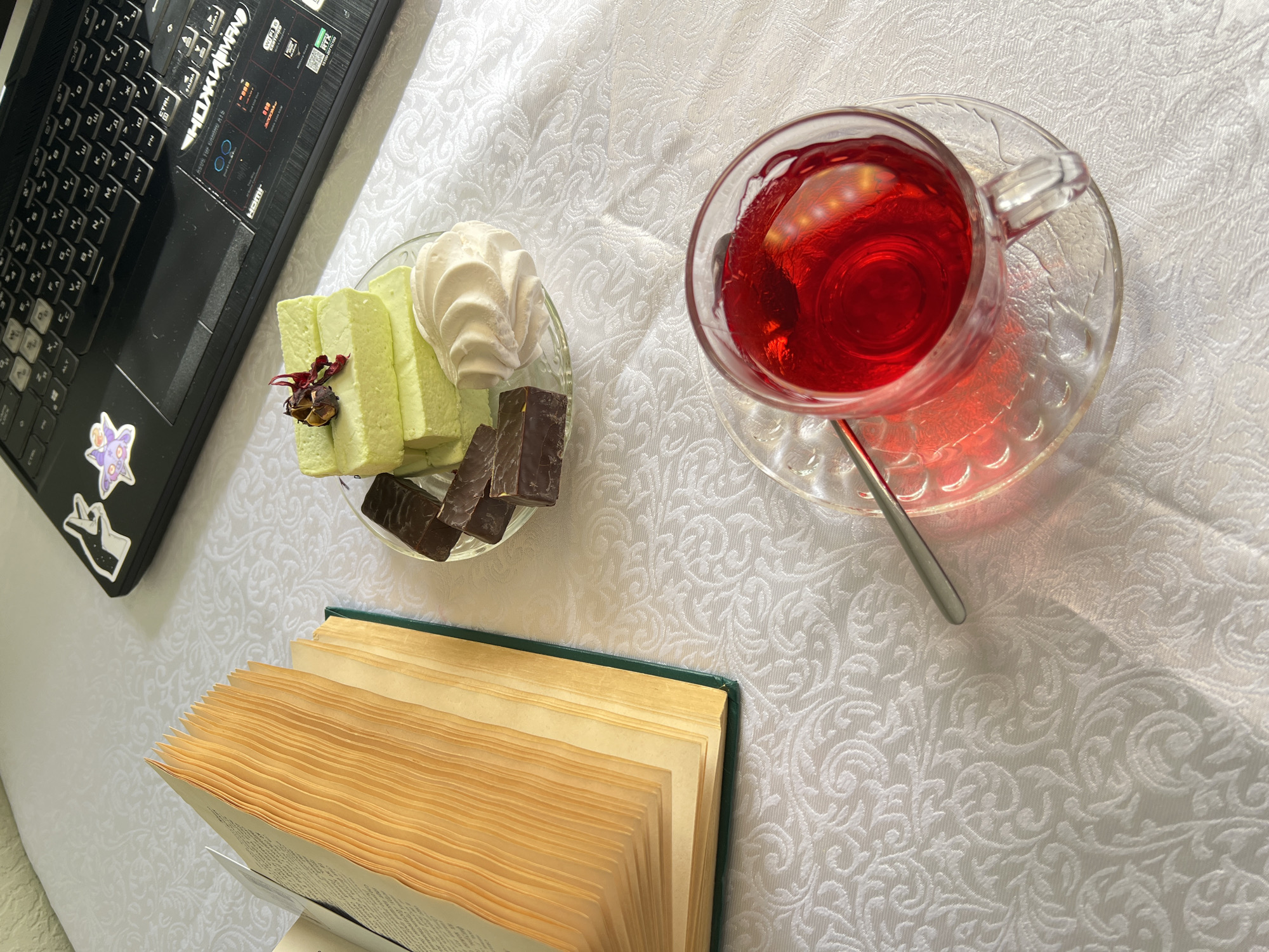 This eye-level, indoor shot depicts a still life arrangement on a white, patterned surface. A portion of a black laptop is visible in the upper left corner, adorned with stickers. Adjacent to the laptop is a small glass plate holding green layered cakes, a white meringue, and pieces of dark chocolate. A clear glass teacup filled with a red liquid and a metal spoon sits to the right. In the lower portion of the image, an open book with yellowed pages and a green cover is positioned, suggesting a reading session. The scene evokes a sense of leisure and relaxation, likely taking place at home or in a cozy café. The setting doesn't inherently link to Saint Petersburg, Russia, as the objects are common and the environment is generic. Additional information is needed to establish the location definitively.