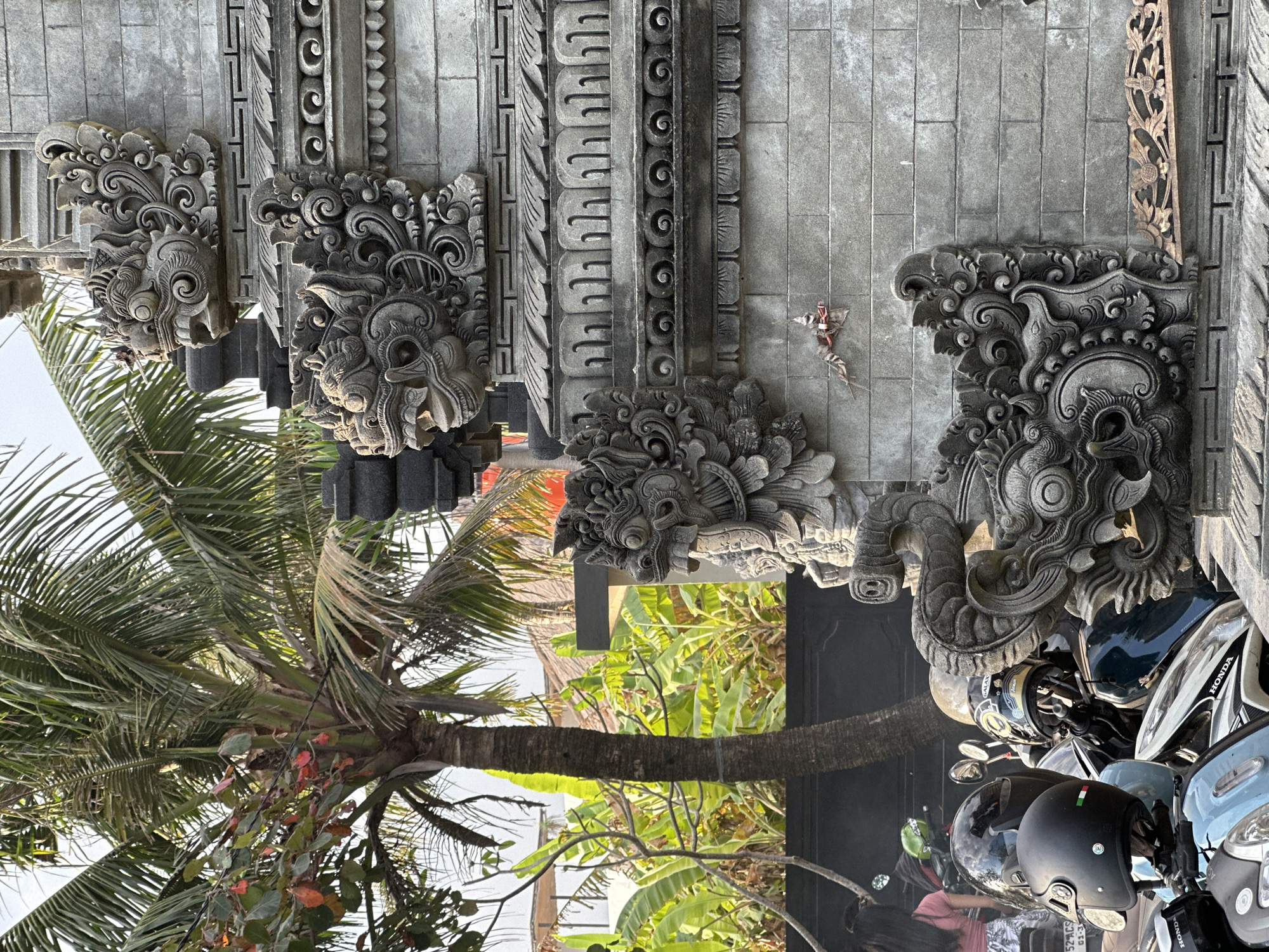 This image, taken in the city of Mengwi, Indonesia, showcases intricate stone carvings that adorn a traditional Balinese temple or gateway. The stonework features detailed mythological figures, likely depicting guardian spirits or deities, with fierce expressions and ornate headdresses. The carvings are set against a backdrop of lush tropical vegetation, including a prominent palm tree and banana plants, suggesting a warm and humid climate. In the foreground, a collection of parked motorcycles, some with helmets, indicates the presence of daily life and transportation in the area, contrasting with the ancient artistry of the stone structures. The scene is bathed in natural daylight, creating distinct shadows and highlighting the textures of the stone and foliage.