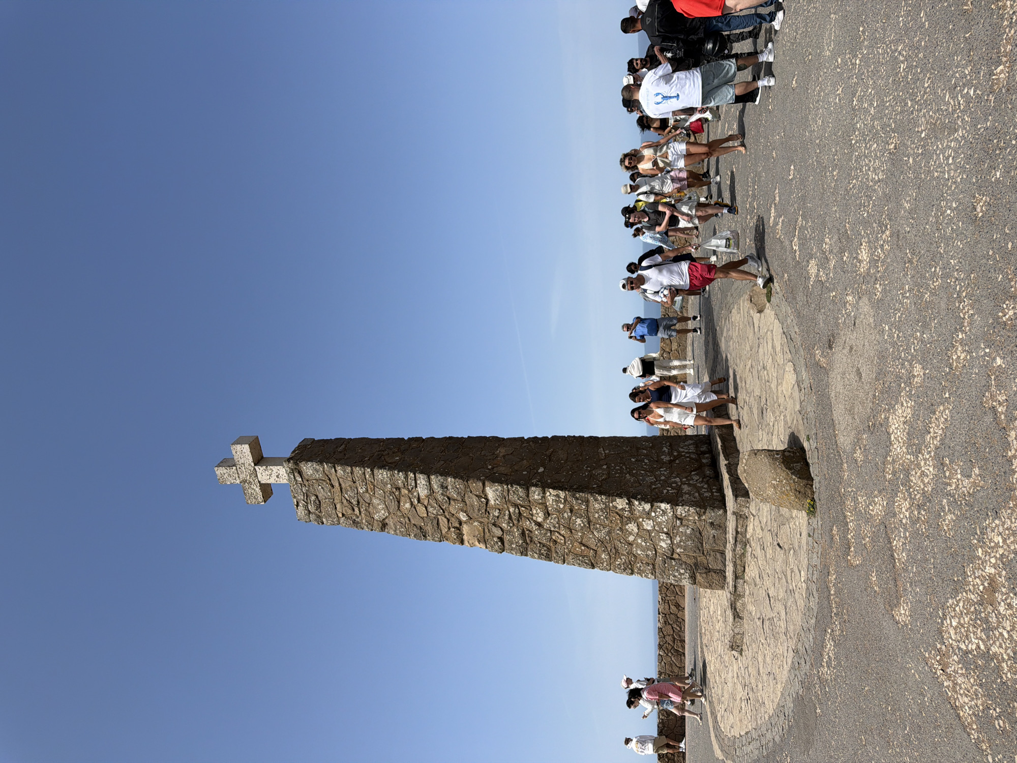 The image depicts a sunny day at Cabo da Roca, the westernmost point of mainland Europe, located in the municipality of Sintra, near Colares, Portugal. A prominent stone monument, a large cross atop a tapering stone pillar, stands in the center-left of the frame against a clear blue sky. The monument is surrounded by a paved area with intricate stonework patterns. A crowd of tourists is present, walking around the monument and enjoying the view. The people are dressed in casual summer clothing, with many wearing shorts, t-shirts, and hats. Some are taking photographs, while others are simply strolling. The environment is coastal, with a hint of the ocean visible in the background, beyond a low stone wall. The overall atmosphere is one of a popular tourist attraction on a bright, clear day.