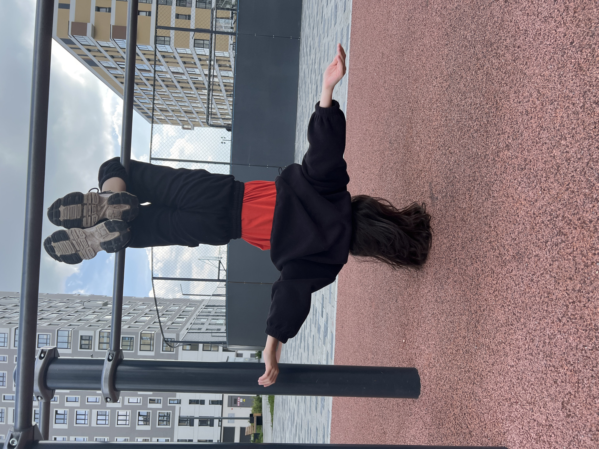 The image depicts an outdoor urban scene featuring an individual performing an inverted hang on a pull-up bar. The setting is a modern residential area within the city of Yekaterinburg, Russia.
In the foreground, a person with long, dark hair is suspended upside down from a horizontal dark grey metal bar. Their head is positioned towards the ground, obscured by their hair. They are wearing a black long-sleeved hoodie or sweatshirt over a red top, black pants, and athletic sneakers with light-colored soles and dark treads. The person's feet and shins are hooked over the horizontal bar, while their arms are extended horizontally outwards from their body, palms facing upwards with fingers slightly curled. The pull-up bar is supported by two vertical dark grey metal poles, connected by visible silver-colored clamps and bolts.
The ground beneath the person and the exercise equipment is covered with a reddish-brown granular, soft-fall material, typical of a playground or sports surface. Beyond this surface, a grey paved area with interlocking pavers is visible.
In the background, several multi-story residential buildings rise against a partly cloudy sky. To the left, a tall, grey apartment building with numerous regular rectangular windows is prominent. To the right, another large apartment building, characterized by its yellow/beige facade with dark-framed windows and balconies, is visible. Between the foreground and the background buildings, a dark metal fence, possibly a chain-link fence, encloses an area, with structures possibly indicating a sports court (like a basketball hoop pole) discernible behind it on the right side. The sky shows a mix of light blue and scattered white and grey clouds, suggesting an overcast but bright day.
