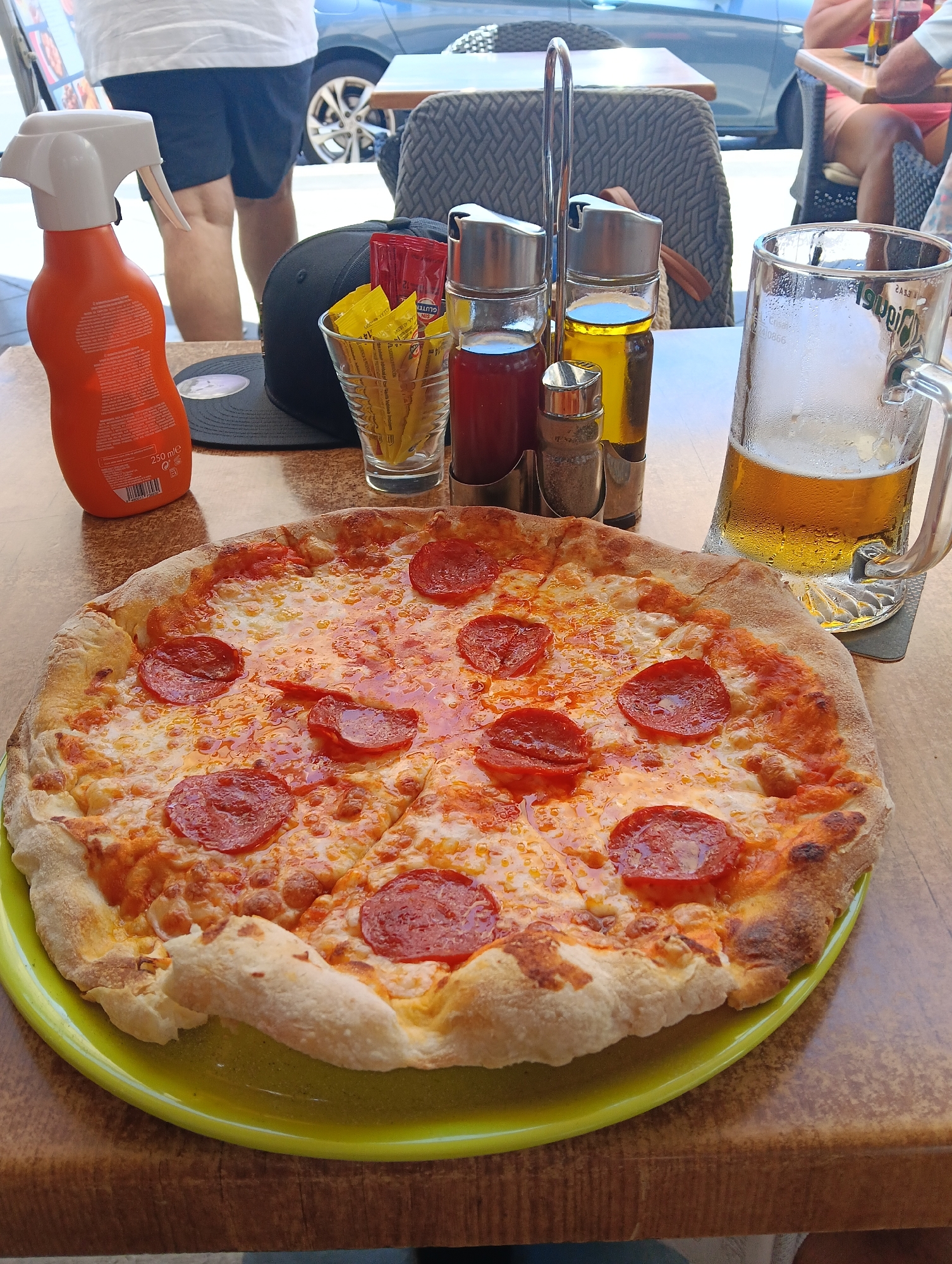 This media file captures a sunny, outdoor dining scene, likely at a restaurant or bar patio in Puerto Del Carmen, Spain. The main focus is a large, freshly baked pepperoni pizza, centrally placed on a bright green plate. The pizza has a golden crust, melted cheese, and evenly distributed slices of pepperoni.
To the right of the pizza, a large glass beer mug, partially filled with amber beer and covered in condensation, rests on a dark coaster, suggesting a cold beverage on a warm day. A silver condiment caddy stands behind the pizza, holding a red bottle (likely ketchup), a bottle of olive oil, and two shakers (possibly salt and pepper). Adjacent to the caddy, a glass contains several individual yellow packets (likely mustard or mayonnaise) and red packets, with "GLUTEN FREE" visible on one red packet.
On the left side of the table, an orange spray bottle, marked with "250 ml e", is present, possibly sunscreen or insect repellent. A black baseball cap lies on the wooden table behind it.
In the background, partial views of two individuals are visible. On the left, a person wearing a white t-shirt and dark shorts is standing. On the far right, another person is seated at an adjacent table. A grey car is parked on a street further in the background. The bright sunlight and outdoor setting suggest a relaxed daytime meal or casual gathering. The beer mug faintly displays the brand "Zipfer" and capacity "0.4L" in reverse.
