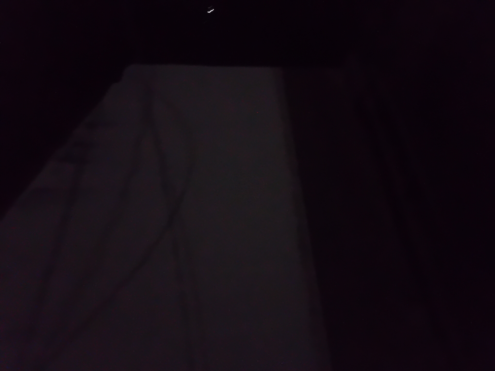 The image depicts a dimly lit scene, predominantly composed of dark tones and shadows. A large, off-white surface occupies the upper portion of the frame, appearing to be a fabric or draped material that is illuminated by an unseen light source. The texture of this surface suggests folds and creases, creating subtle variations in shade. The lower portion of the image is almost entirely black, indicating a dark environment or a deep shadow cast by an object. In the far right of the frame, there is a bright, curved white line, possibly a reflection or a distant light. There are also a few very faint red and white specks scattered across the image, which could be dust, light reflections, or distant points of light. The scene's context within Jauro Yinu, Nigeria, is not evident from the visual information presented in the image itself.