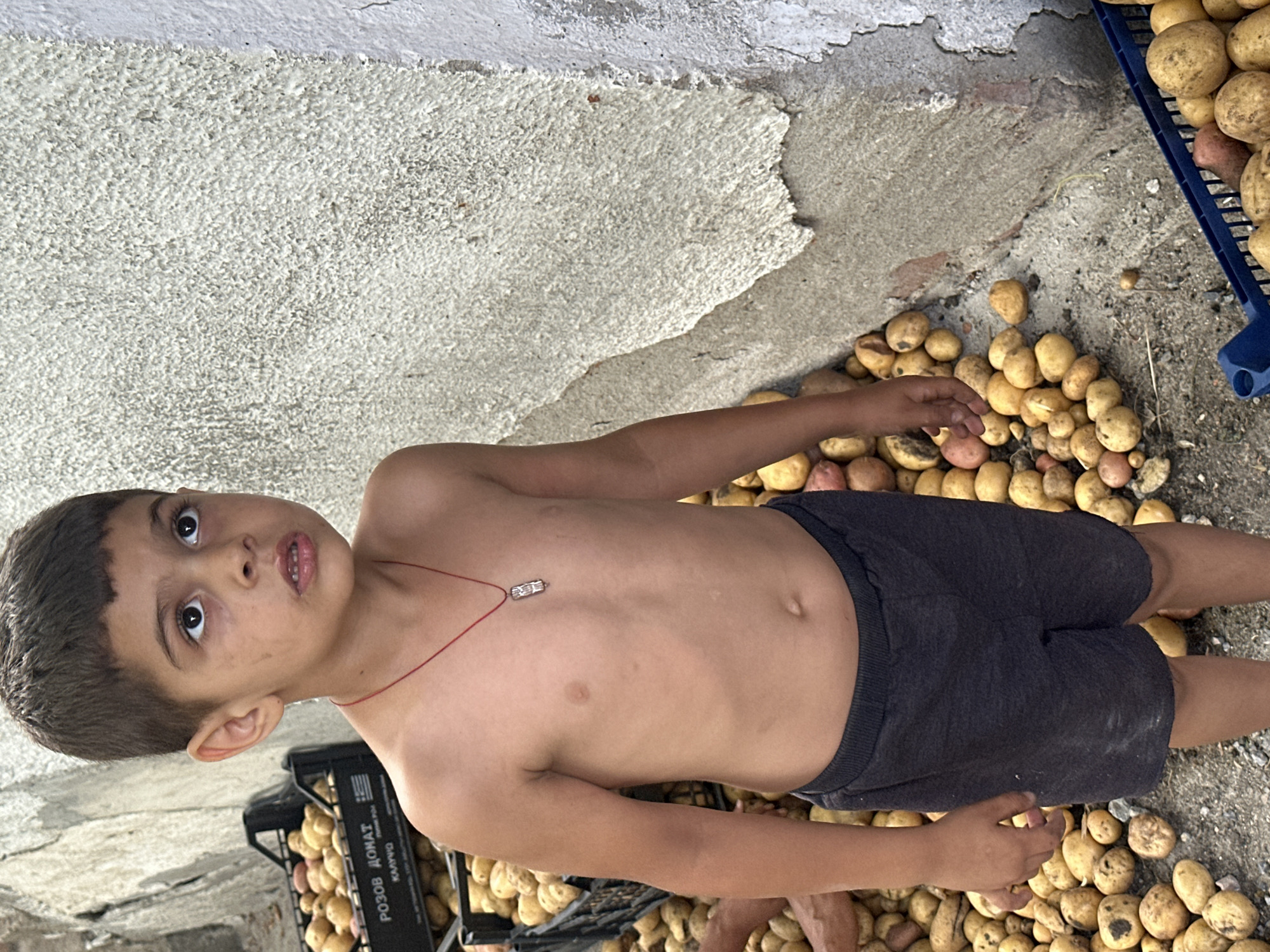 A young boy stands in front of a textured, light-colored wall in Mlekarevo, Bulgaria, looking upwards and to the right with a slightly open mouth. He is shirtless, wearing dark shorts, and a red string with a rectangular pendant around his neck. He is surrounded by potatoes. To his left, a black plastic crate is filled with potatoes, with text in a foreign language on its side. More potatoes are scattered on the ground around him, with a blue plastic crate visible in the lower right corner, also containing potatoes. The ground appears to be gravel and dirt. The lighting suggests it is daytime.