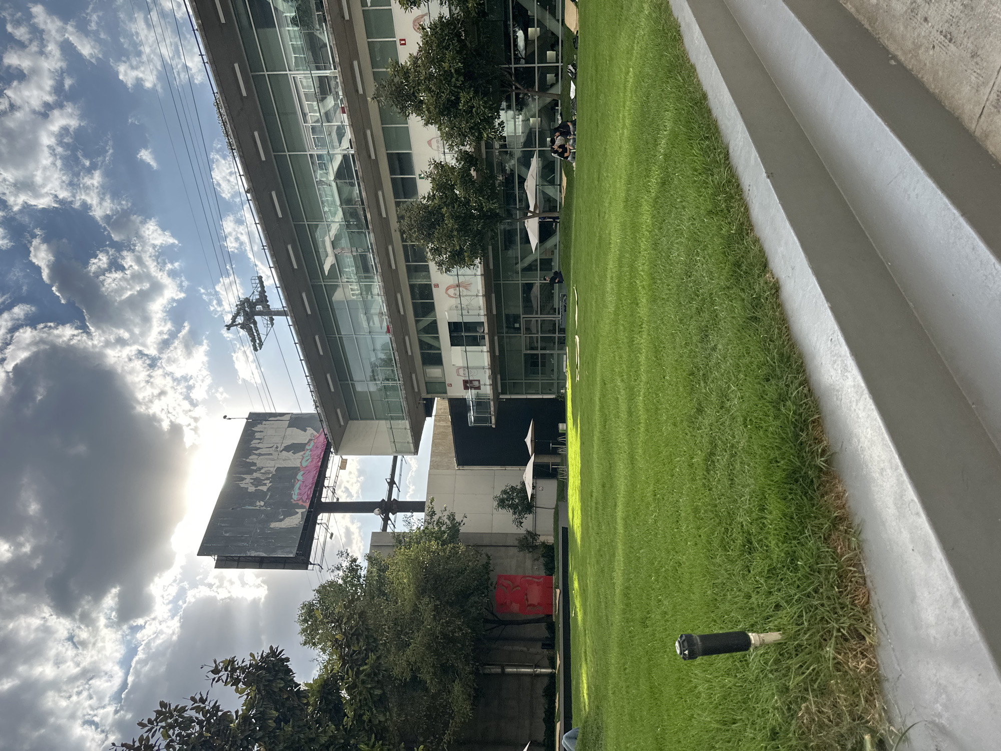 This image captures a vibrant urban outdoor scene in Mexico City, Mexico, on a bright, partly cloudy day, likely in the late afternoon given the quality of sunlight. The scene features a modern glass-fronted building, a lush green lawn, and elements of a cable car system.
In the foreground, a neatly trimmed green grass lawn occupies the left and center, bordered on the right by grey concrete steps. A black sprinkler head is visible in the grass near the bottom right. In the middle ground, several people are seen relaxing or socializing at tables under white umbrellas on a paved area adjacent to the building. Various trees with green foliage are interspersed throughout, creating shaded spots.
To the left, a large, weathered billboard with faded, colorful artwork (possibly graffiti or street art, but no legible text) stands prominently against the bright sky, partially backlit by the sun. Below the billboard and amidst trees and concrete structures, a distinct, abstract red sculpture is visible.
The background is dominated by a multi-story, modern building with extensive glass windows that reflect the sky. Some subtle wall art, appearing as outlines of faces, can be seen on a lower section of the building's facade. Above the building and billboard, against a sky with scattered white and grey clouds, a cable car pylon and its suspension lines are clearly visible, serving as a strong location cue for Mexico City's Cablebús system. The overall atmosphere suggests a leisurely urban environment integrating green spaces with modern architecture.