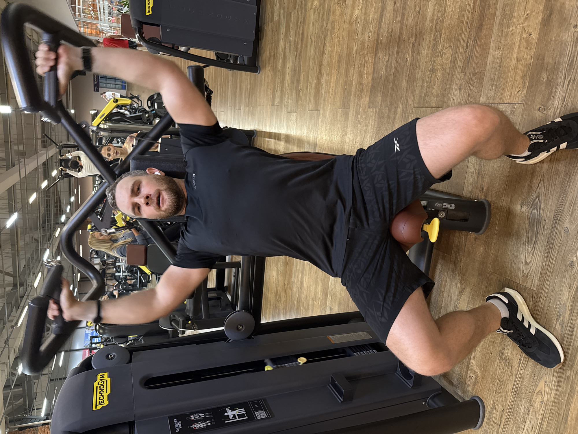 A man is using a Technogym shoulder press machine in a gym. The man is wearing a black t-shirt and black patterned shorts, with black and white sneakers. The gym has a wooden floor and other gym equipment visible in the background. The location is Chaikovsky, Russia.
