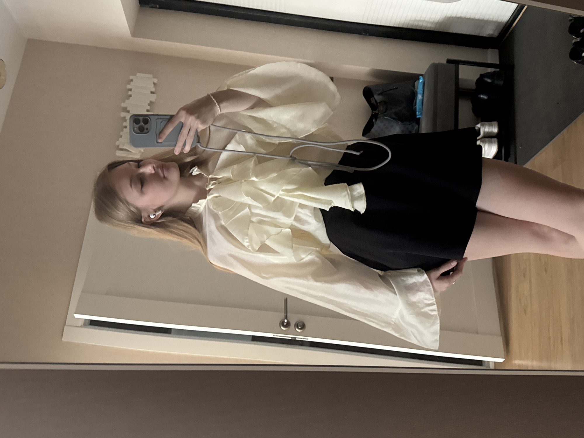 A young woman with blonde hair, wearing a cream-colored ruffled blouse and a black skirt, takes a mirror selfie in what appears to be a residential interior, possibly an apartment in Perm, Russia. She is holding a smartphone in her right hand, angled to capture her reflection. She has white earbuds in her ears and a thin bracelet on her left wrist. The background shows a doorway with a silver handle, a beige wall with a decorative wall sculpture, and a grey bench with a dark bag and some shoes on the floor behind her. The flooring is a light wood laminate. The overall impression is of a casual personal moment captured at home.