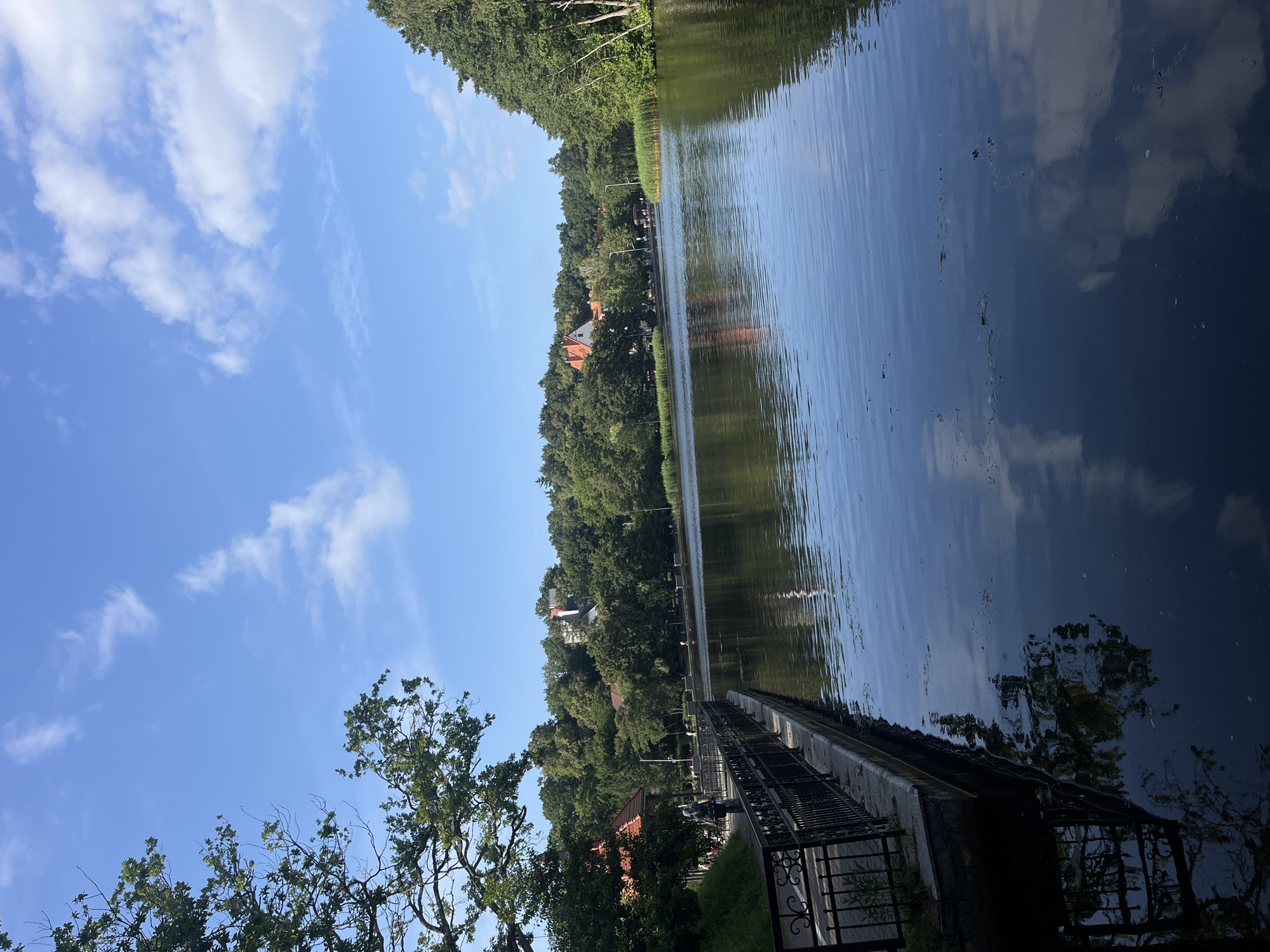 The image depicts a calm body of water, likely a lake or river, reflecting the sky and surrounding environment. On the left side of the image, a paved walkway runs along the water's edge, bordered by a black metal railing. Beyond the walkway, there are trees and buildings with red roofs, suggesting an urban or park setting. Several people are visible walking or cycling on the path. The water itself appears dark and still, with subtle ripples and some floating debris. The sky is blue with scattered white clouds. The overall scene conveys a peaceful atmosphere, characteristic of a recreational area in a city like Svetlogorsk, Russia, possibly the city's embankment or a park area adjacent to a body of water.
