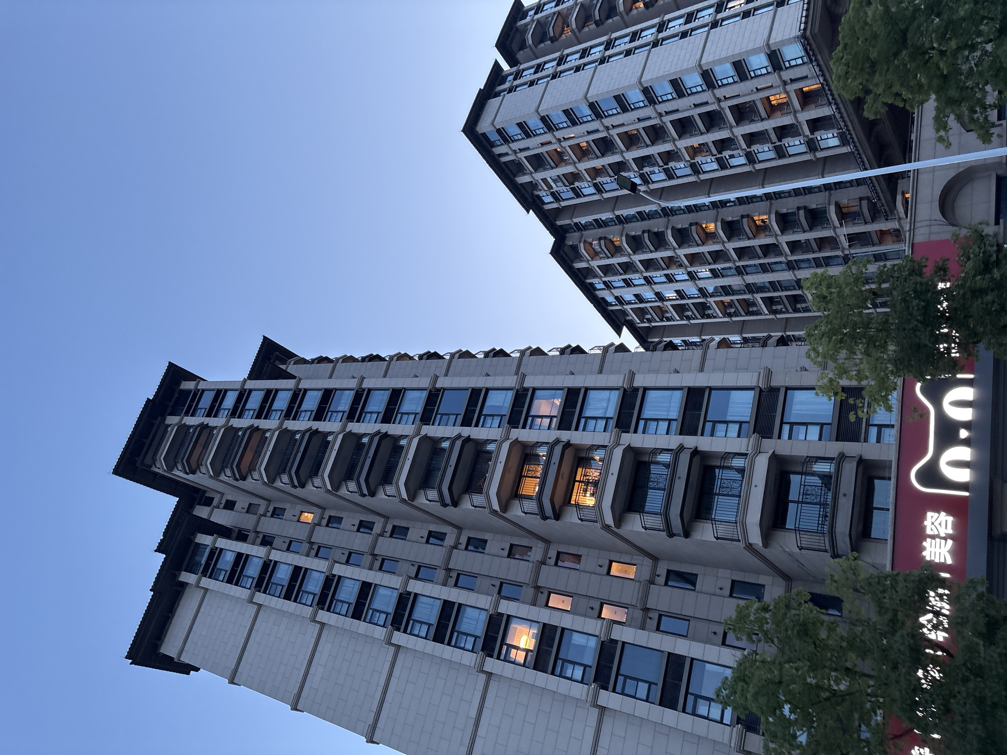 In the Minhang District of China, two tall, modern apartment buildings rise against a clear blue twilight sky. The buildings are constructed with light-colored stone facades and feature numerous windows and balconies. Some windows are illuminated, suggesting that residents are present within the apartments. The architecture is characterized by sharp angles and clean lines, with dark-colored roof overhangs. In the foreground, the tops of green trees are visible, and a brightly lit red sign with white lettering and a cartoon cat logo is partially obscured by the foliage. A tall, white lamppost stands between the two buildings, extending upwards into the sky. The overall scene depicts a residential area in an urban environment during the evening.