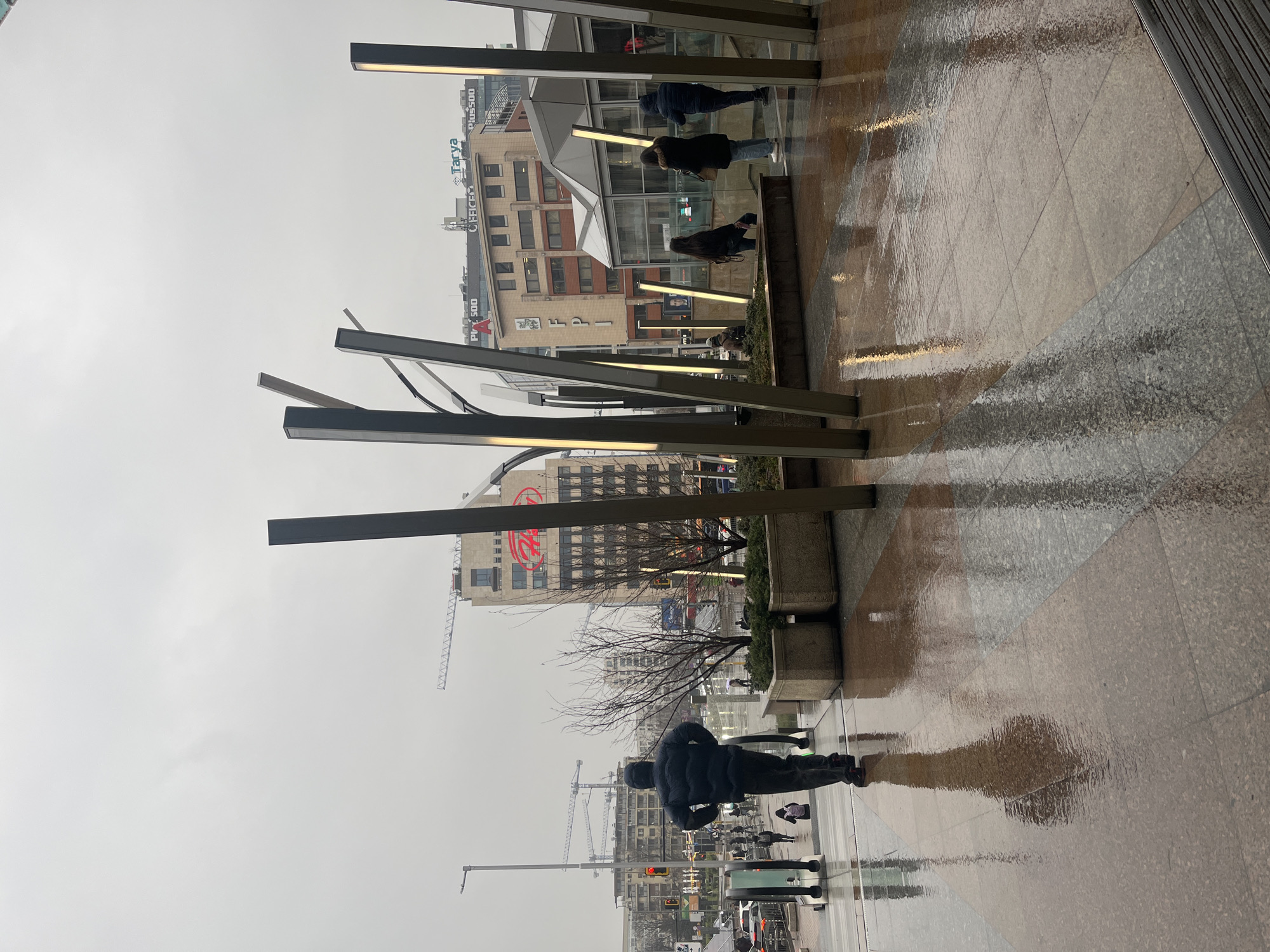 The image depicts an outdoor scene in Sofia, Bulgaria, on a rainy day. The foreground shows a section of wet, tiled pavement reflecting the surroundings. A series of modern, rectangular lighting fixtures stand along the edge of the pavement, some angled. People are walking along a path behind the lighting, visible through the gaps between them. In the background, buildings are visible, including one with a prominent red "FPI" logo. Construction cranes are also present, suggesting ongoing development in the area. The sky is overcast, contributing to the overall gray and wet atmosphere.