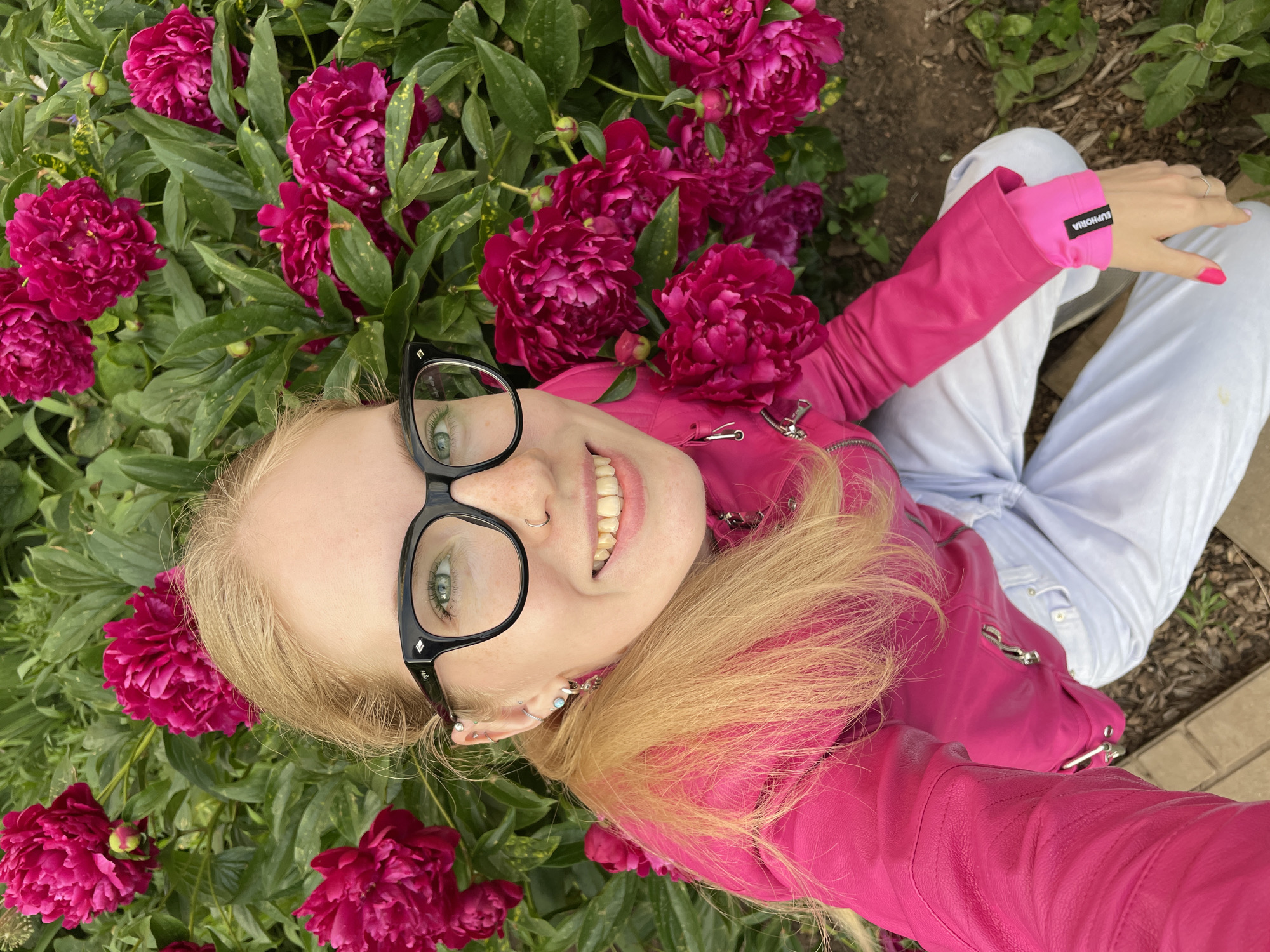 A young Caucasian woman with blonde hair and green eyes is taking a selfie in front of a bush of pink peonies. She is wearing a bright pink jacket, white pants, and black-rimmed glasses. She has several piercings in her ears and nose. The peonies are in full bloom, with many large, dark pink flowers and green leaves. The background is a garden, with some dirt and leaves visible on the ground. The woman appears to be happy and smiling. There is no specific information that can be used to determine the location in Izhevsk, Russia.