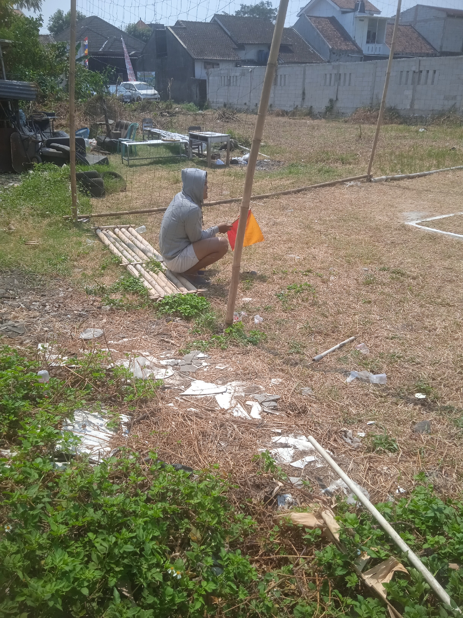 In Klodran, Indonesia, a person wearing a grey hooded sweatshirt and white shorts is squatting next to a bamboo mat on a dry, grassy field. The person is holding a red and yellow flag. The surrounding environment is cluttered with debris, including plastic bottles, white paper scraps, and dried vegetation. In the background, there are houses with tiled roofs, a concrete wall, and a soccer net. A white pole is visible in the foreground. The scene appears to be an outdoor area that may be used for sports or recreation, but it is currently unkempt and littered.