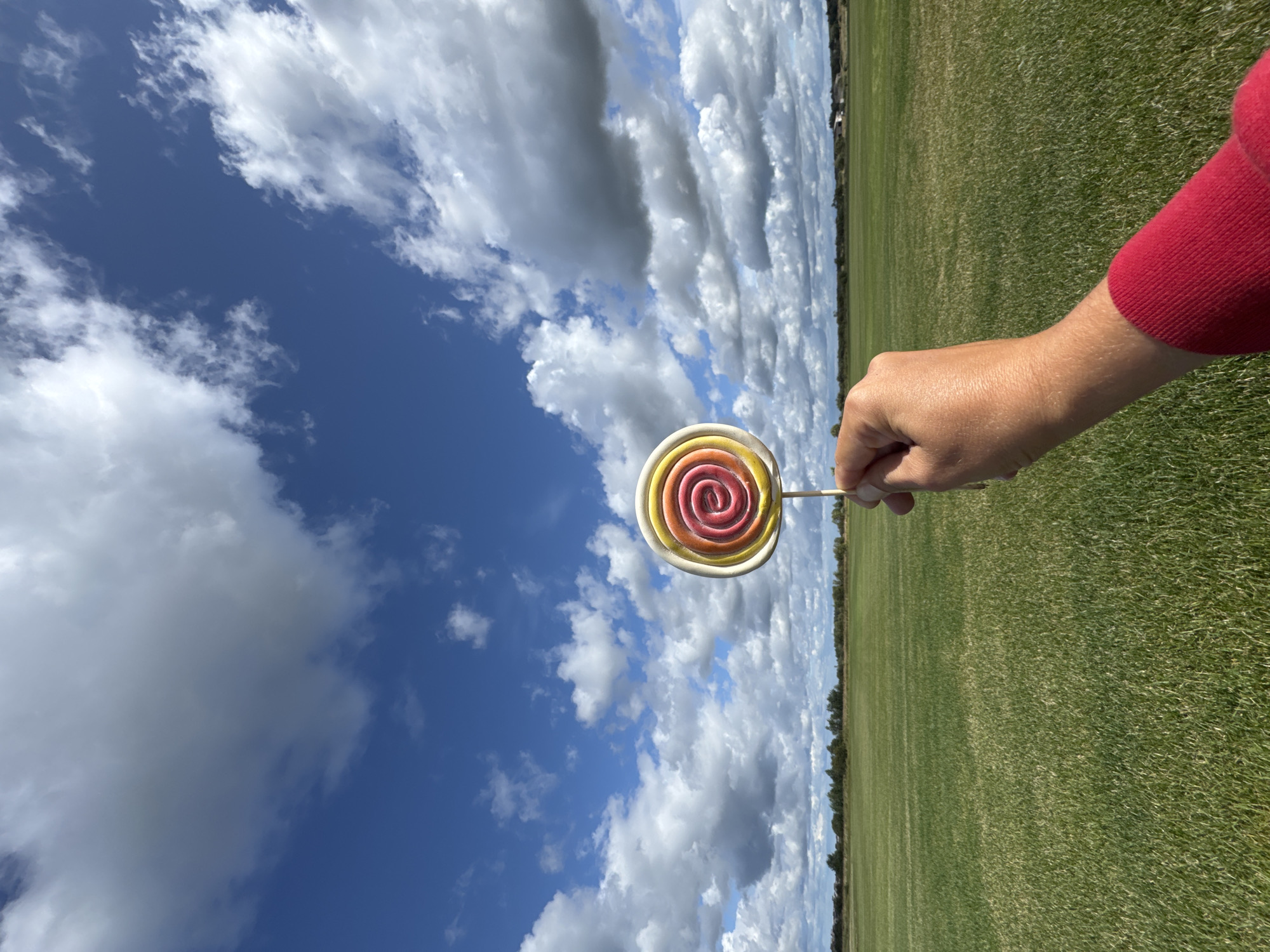 In Kempelevo, Russia, a person's hand, adorned with a red sleeve, holds a colorful, spiral lollipop against a backdrop of a vast, green field under a dynamic, cloudy sky. The lollipop exhibits concentric circles of red, yellow, and white. The field stretches to the horizon, where a line of trees and distant structures are faintly visible. The sky is a vivid blue, punctuated by large, white, and grey cumulus clouds, suggesting fair weather with potential for passing showers. The scene is captured from a low angle, emphasizing the expanse of the sky and the bright lollipop.