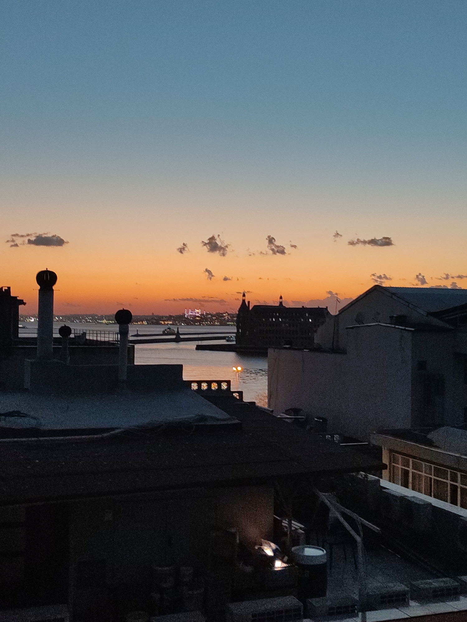 The image captures a sunset over the Bosphorus Strait, as viewed from the rooftops of Kadıköy, Turkey. The sky transitions from a deep blue at the top to a vibrant orange and pink hue near the horizon. Silhouetted against this colorful sky are the distinct architecture of buildings and structures within Kadıköy, including prominent chimney stacks on the foreground rooftops. In the middle ground, the water of the strait is visible, with various vessels including ferries and boats navigating the waterway. The distinctive Haydarpaşa Terminal building stands out on the distant shore, its silhouette dark against the sunset. The foreground consists of various rooftop structures, some appearing to be residential or commercial buildings with visible ventilation systems and antennae. The lighting suggests it is either early evening or late afternoon, with artificial lights beginning to illuminate some of the structures. There are no discernible people or animals in the frame.