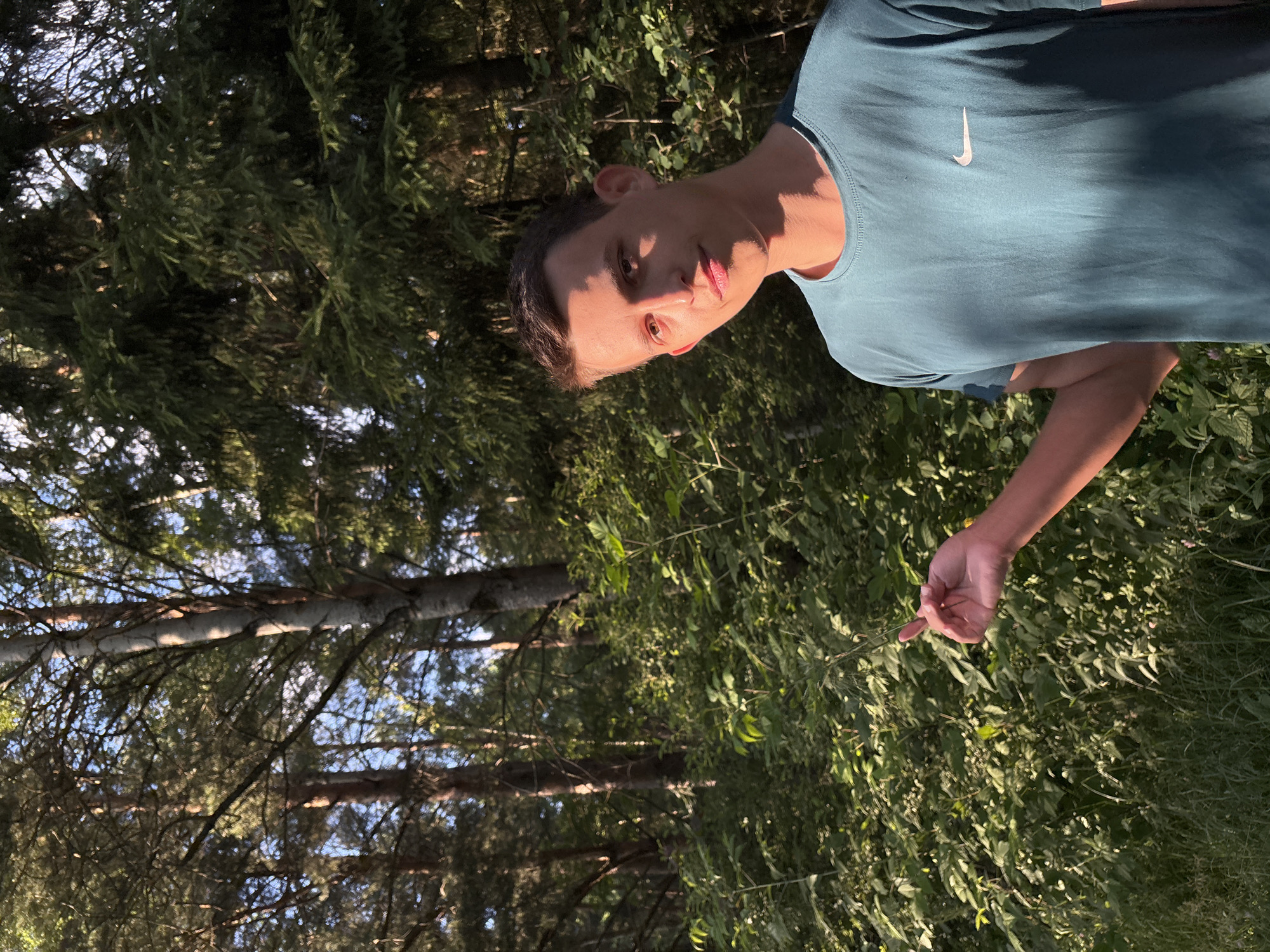 A young man is pictured in a wooded area in Shevl'yagino, Russia. He is wearing a teal t-shirt with a white Nike logo on the left chest. The man is looking towards the camera with a neutral expression, and sunlight is casting a shadow across his face. He is holding a thin stem of a plant in his right hand. The background is filled with lush green foliage and trees, suggesting it is daytime and likely summer or early autumn. The lighting indicates it is either morning or late afternoon, with the sun creating warm highlights and shadows. The overall impression is one of a casual outdoor moment. No visible text other than the Nike logo.