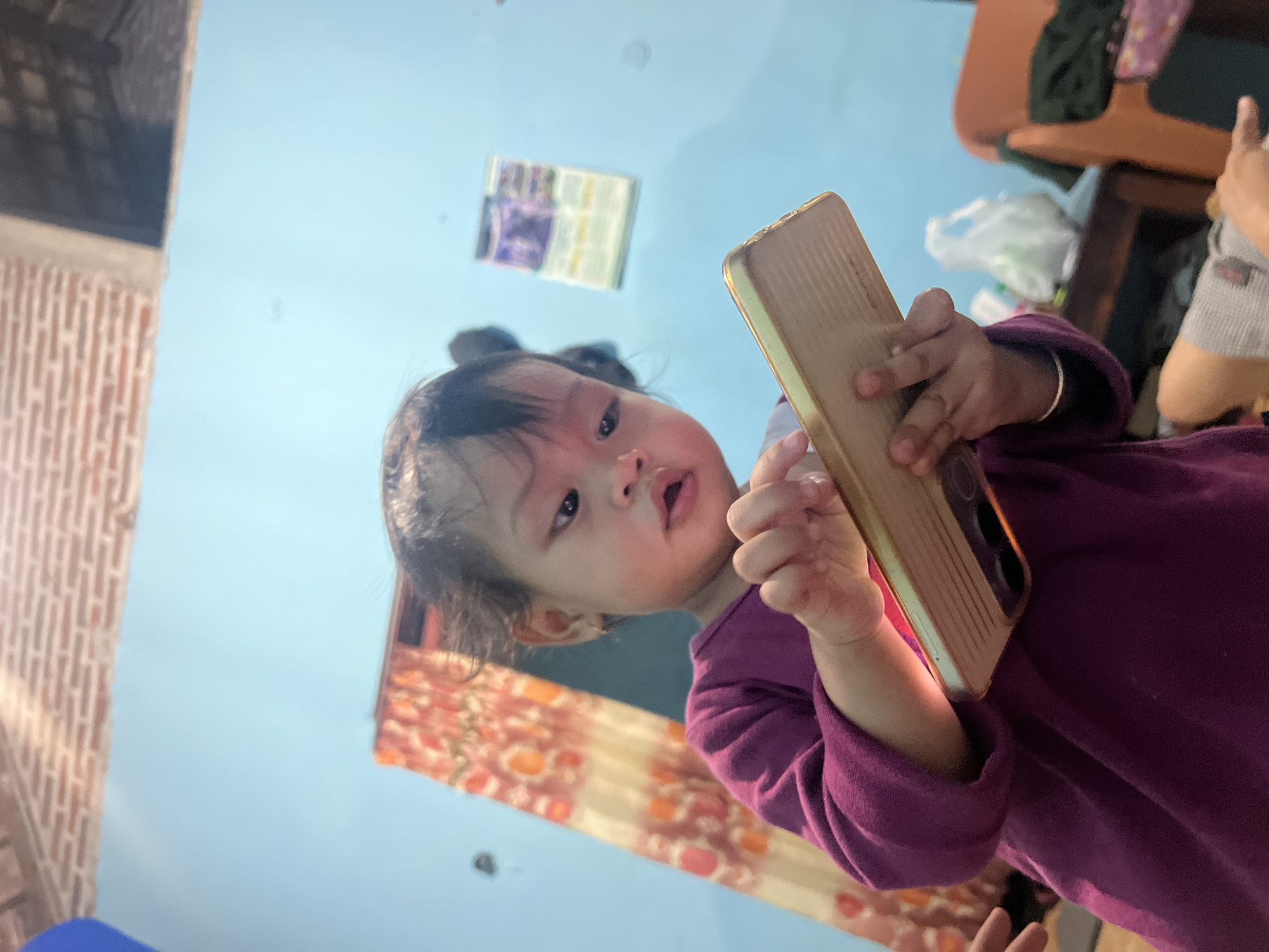 This image, taken in Kecamatan Pati, Indonesia, captures a close-up, low-angle perspective of a young child, likely a toddler, holding a smartphone. The child, with dark hair, wears a purple long-sleeved top or dress and looks directly at the camera with a neutral to slightly curious expression, lips slightly parted. They are holding a smartphone with a distinctive gold-colored, ribbed case.
The indoor setting features a dominant light blue wall in the background. To the left, a section of a red brick-patterned wall or structure is visible. Below that, and behind the child, patterned curtains with an orange and brown floral or abstract design are present. A rectangular object, possibly a framed picture or calendar, hangs on the light blue wall in the upper background. To the right, a part of another person's arm or hand, covered in a grey sleeve, is visible. Further in the upper right corner, hints of household furniture, such as a chair or table with a white plastic bag and dark fabric on it, can be seen. The bright illumination suggests it is daytime. There is no visible text in the image. The scene conveys a casual, everyday moment of a child interacting with a mobile device within a home environment.