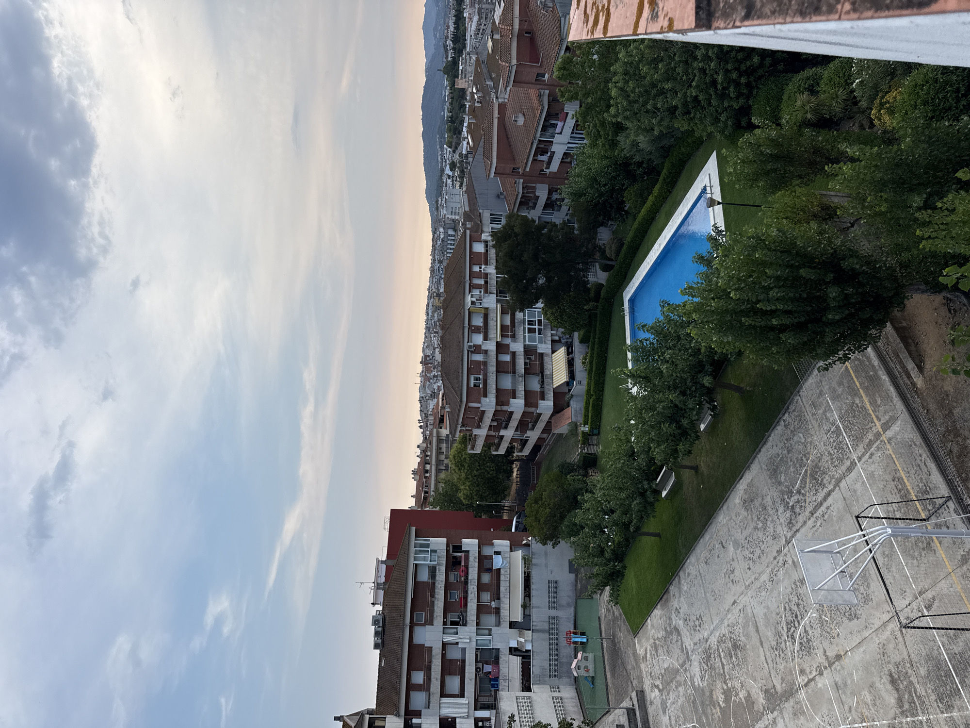 This is a high-angle, medium shot of a residential area in Blanes, Spain. The image captures a collection of apartment buildings with red tile roofs and white balconies. In the foreground, a concrete basketball court with faded white lines and a goal is visible. Adjacent to the court is a patch of grass with several trees and two white benches. Further back, a bright blue swimming pool with a white border is situated within a manicured lawn area. The scene is bordered by lush green trees and foliage. In the background, a sprawling cityscape with numerous buildings extends towards a distant, hazy mountain range under a cloudy sky. The overall impression is one of a tranquil, suburban neighborhood.