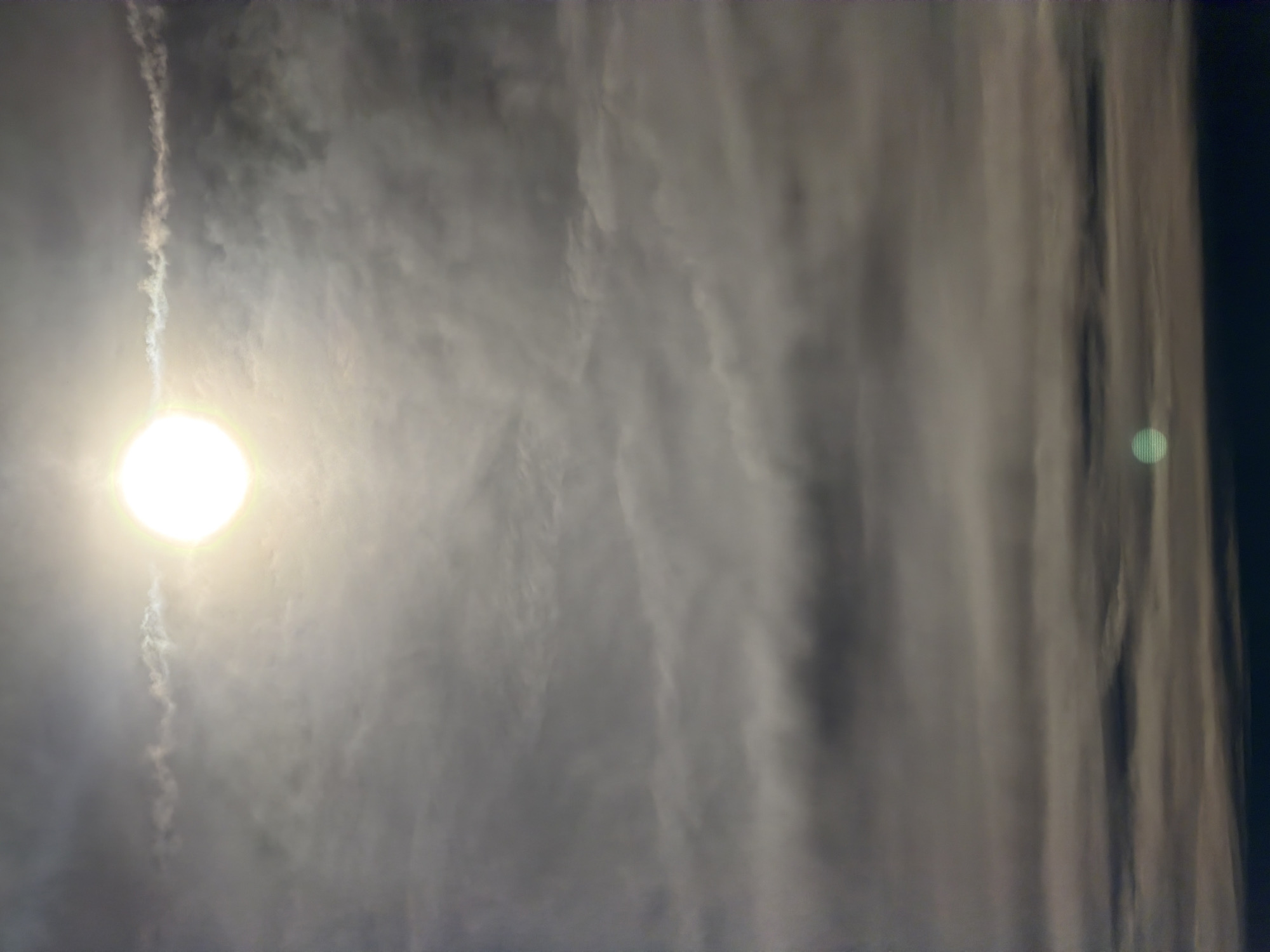 The image shows a sky with the sun obscured by clouds, appearing as a bright, hazy orb. Below the sun, layers of gray clouds stretch across the frame, with the density and texture varying. A faint, greenish circular anomaly is visible in the lower portion of the image. The bottom edge of the image appears to be the dark horizon, suggesting a landscape or cityscape below, but these elements are not clearly discernible. The overall impression is of an overcast day with the sun attempting to break through the clouds.