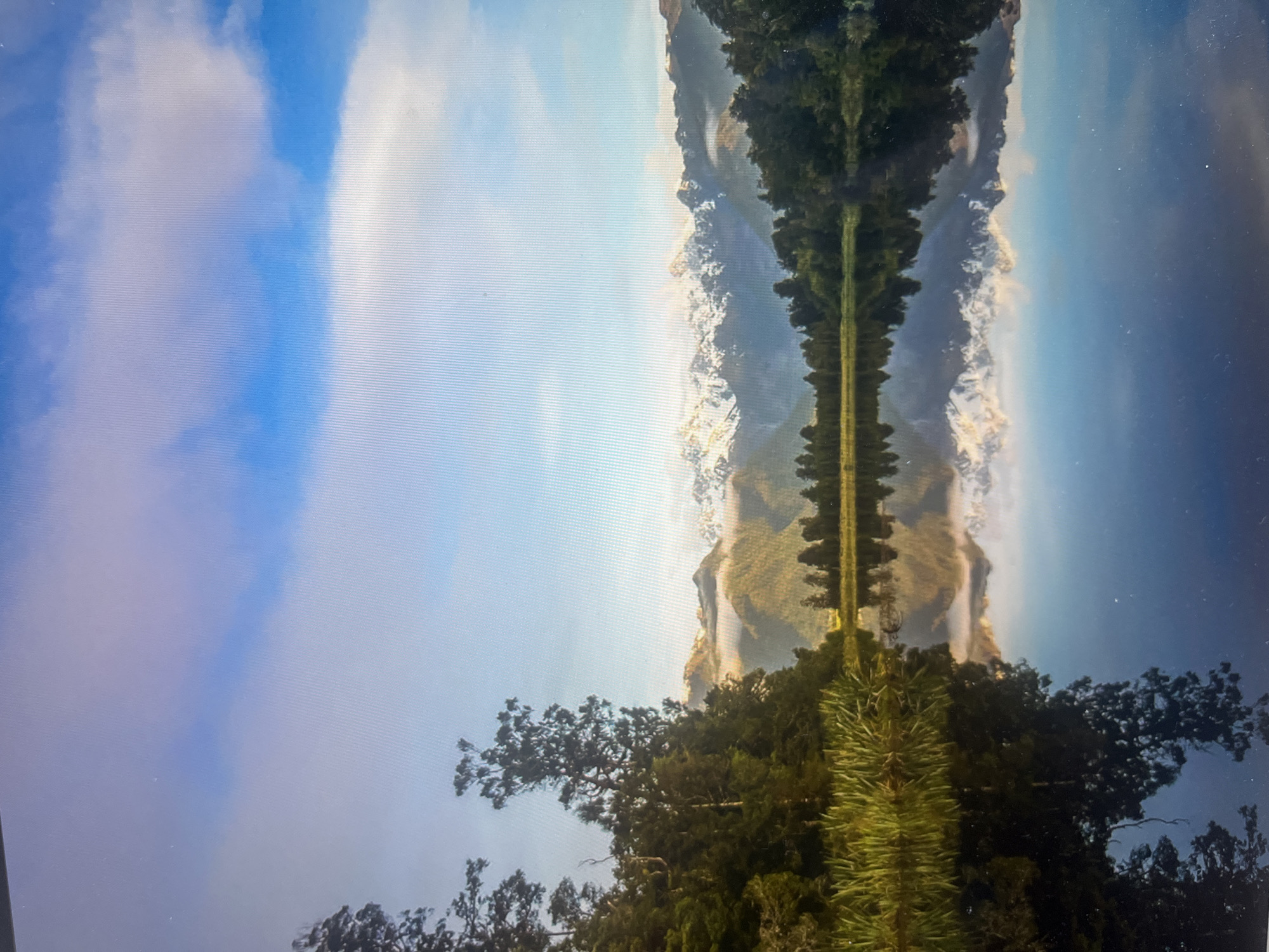 The image is a portrait-oriented photograph featuring a natural landscape heavily manipulated with a horizontal mirroring effect.
**Subjects:** The scene depicts a vast sky with white and grey clouds on the left side of the frame. In the center, there is a prominent range of rugged mountains, some appearing snow-capped or brightly lit, flanked by dense dark green coniferous trees. A striking, narrow, bright green, textured strip runs horizontally across the middle of the image. The right side of the image shows a darker blue expanse, potentially a body of water or a darker sky.
**Activity or event:** No specific activity is depicted; it is a static landscape composition.
**Notable details:** The most dominant feature is the strong horizontal symmetry created by a mirroring effect. The upper portion of the central landscape elements (mountains, trees, and parts of the sky) is reflected in the lower portion of the image, with the bright green strip acting as the approximate axis of this reflection. This manipulation gives the image an abstract and dreamlike quality. The photograph appears to have been taken of a digital screen, evidenced by visible horizontal lines and subtle glare. The bright blue sky and lighting suggest it is daytime. No specific geographical landmarks visible directly indicate the given location context of Ulyanovsk, Russia, due to the abstract nature of the image.
**Visible text:** There is no visible text.
