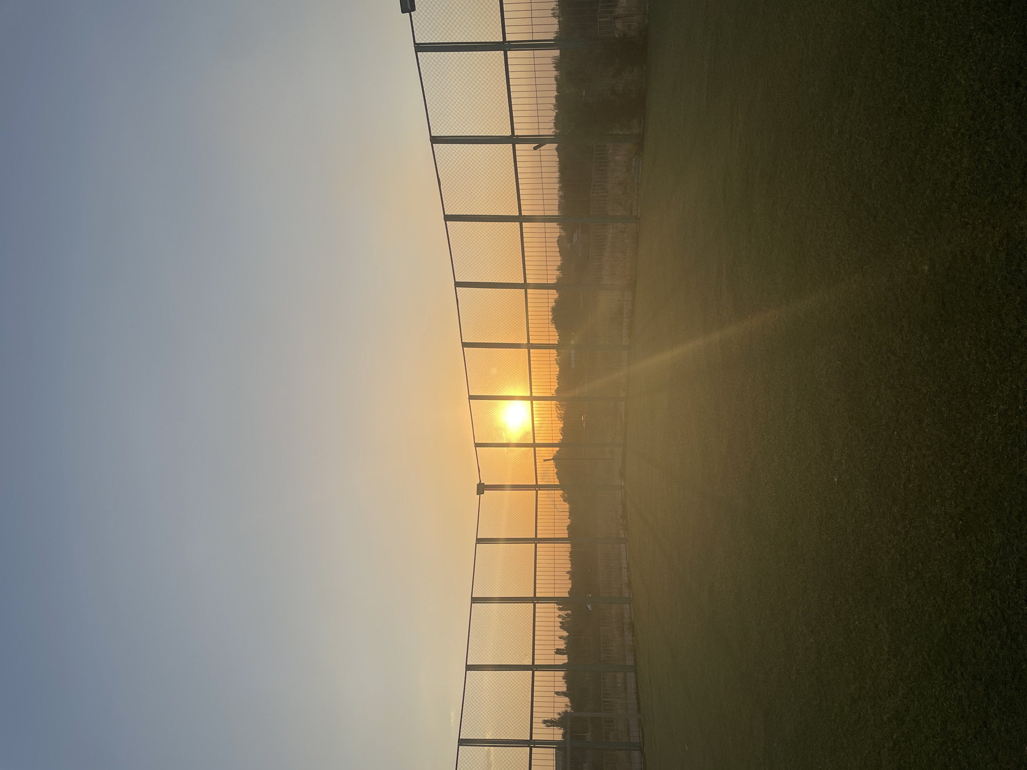 The image depicts a sunset over a fenced, grassy field, possibly a sports field, in Almaty, Kazakhstan. The sun is low on the horizon, casting a warm, golden glow and long shadows across the grass. A chain-link fence with vertical supports runs across the middle ground, separating the field from a background landscape of trees and buildings. The sky transitions from a pale blue at the top to a bright orange and yellow near the sun. There are no people or animals visible in the image. The scene has a tranquil and serene atmosphere.