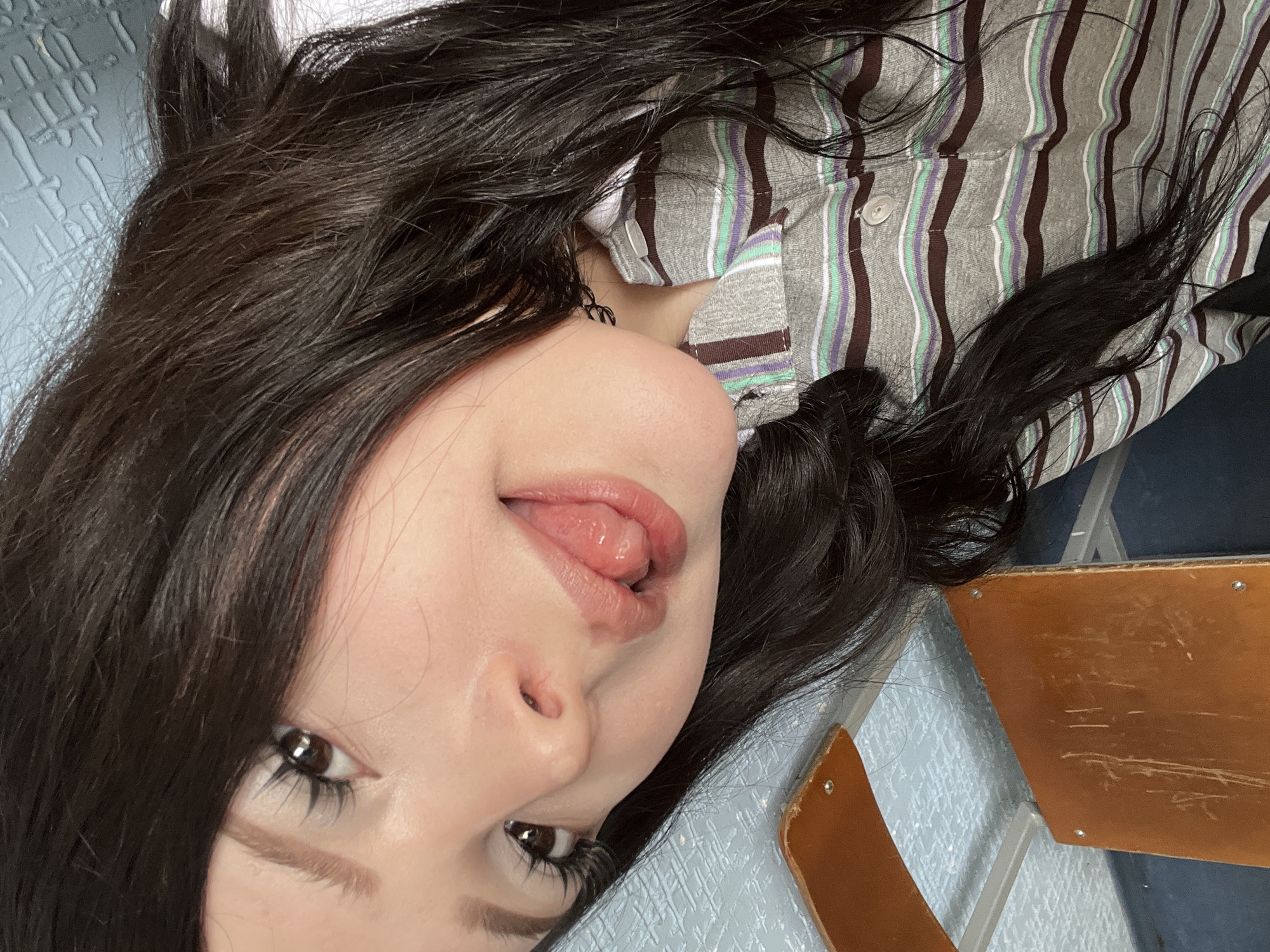 The image shows a close-up, slightly tilted selfie of a young woman with long, dark hair. She is wearing a gray polo shirt with horizontal stripes of brown, purple, and light green. Her tongue is sticking out, and she has long eyelashes. The background appears to be an indoor setting with a textured blue surface and parts of what looks like wooden furniture. There are no visible elements in the image that would allow for the determination of its location in Sterlitamak, Russia.