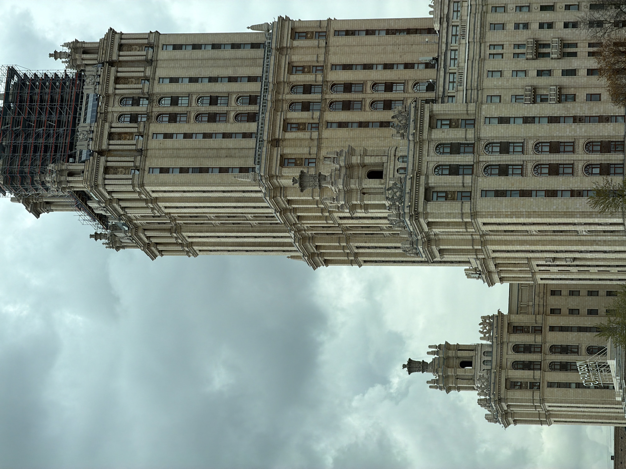 The image displays an urban scene in Moscow, Russia, featuring two grand architectural structures under an overcast sky. The primary subject is a towering, multi-story building constructed in the Stalinist Empire style, characterized by its light-colored stone facade, intricate decorative elements, pilasters, and a mix of arched and rectangular windows. A significant portion of the upper left side of this main building is covered in extensive black scaffolding, indicating ongoing construction or renovation.
A second, smaller building, also exhibiting similar ornate architectural details, is visible to the right and slightly lower. This structure features multiple levels with windows and a decorated pinnacle. A white vertical sign with black text, prominently displaying "RADISSON COLLECTION," is mounted on its facade, identifying it as the Radisson Collection Hotel, Moscow (formerly Hotel Ukraina).
The sky is a uniform, heavy grey, filled with clouds, suggesting an overcast day with no direct sunlight. Sparse, bare tree branches are visible near the buildings, implying the photo was taken during late autumn or winter. The overall atmosphere is stately and emphasizes the grandeur of the architecture against the muted sky. No human activity or specific event is captured; the image is a static architectural study.