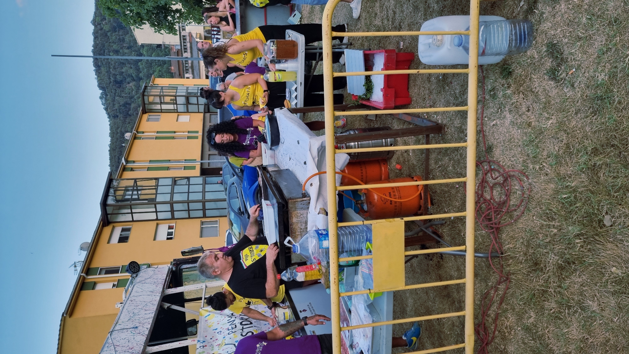 The image depicts an outdoor scene in the city of Sant Joan Les Fonts, Spain, with a group of people gathered around tables laden with food and drinks. In the foreground, a yellow metal barricade separates the viewer from the activity. Behind the barricade, two orange propane gas cylinders are visible, one with a red "REPSOL" logo. A white plastic water container and a large clear plastic bottle filled with water are also present on the ground.
The central focus is a group of individuals, some wearing yellow shirts with the word "STOKATS" printed on them, suggesting a team or organization. A man in a black t-shirt with a similar logo is operating a large griddle, cooking food. A woman with dark, curly hair is standing next to him, looking down at the griddle. Another woman in a yellow "STOKATS" shirt is holding a small food item, possibly a pastry or burger, and looking at it. A third woman in a yellow shirt is also present, with a large pitcher of dark liquid on the table in front of her. There are bottles of water and possibly other beverages on the tables. In the background, a yellow building with green accents and balconies is visible, along with parked cars and a wooded hillside. The ground is covered in dry grass, indicating a warm climate. The overall impression is of an outdoor event or food stand, possibly a community gathering or festival.