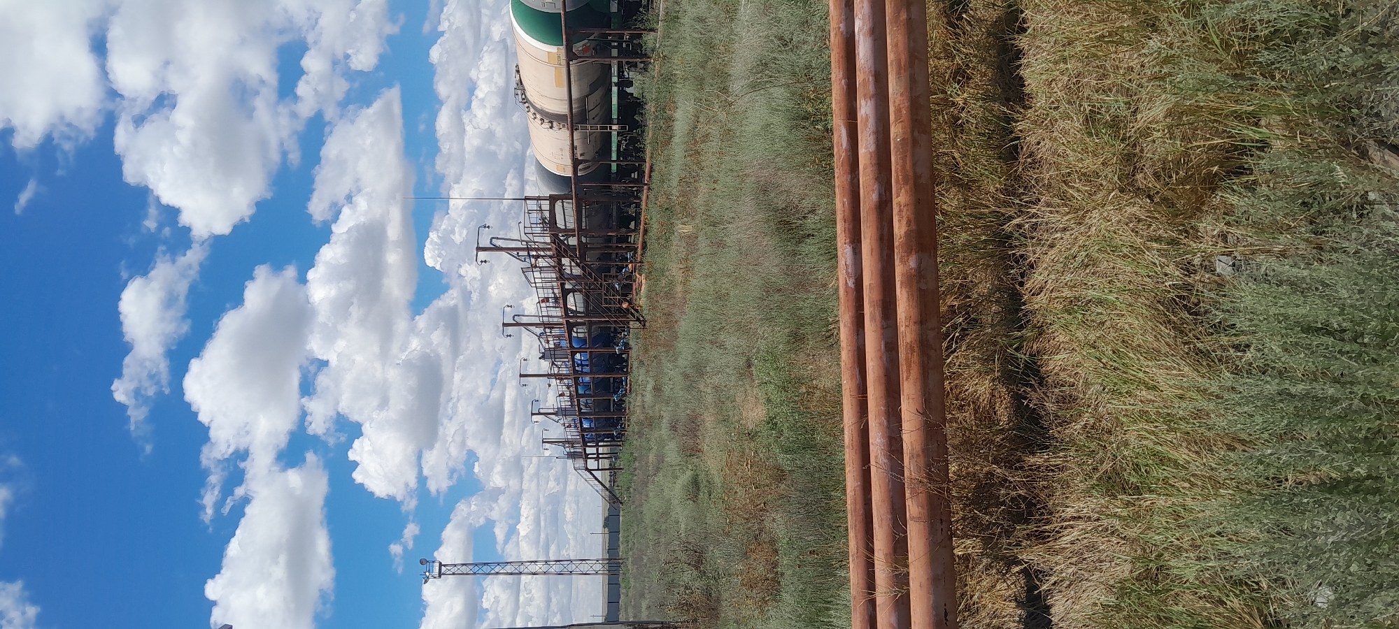This is a medium shot of an industrial area with oil or gas transport infrastructure, located in the city of Embi, Kazakhstan. The scene is dominated by large, rust-colored pipes in the foreground, lying horizontally in the dry, grassy terrain. In the background, a network of elevated metal structures, likely part of a loading or unloading facility, supports multiple tanker cars. These cars appear to be either blue or white and green. A tall, metal tower with a lighting fixture is visible to the left, adding to the industrial feel. The sky above is a bright blue, dotted with fluffy white clouds. The overall impression is one of arid, industrial landscape under a partly cloudy sky. There are no people visible in the image.